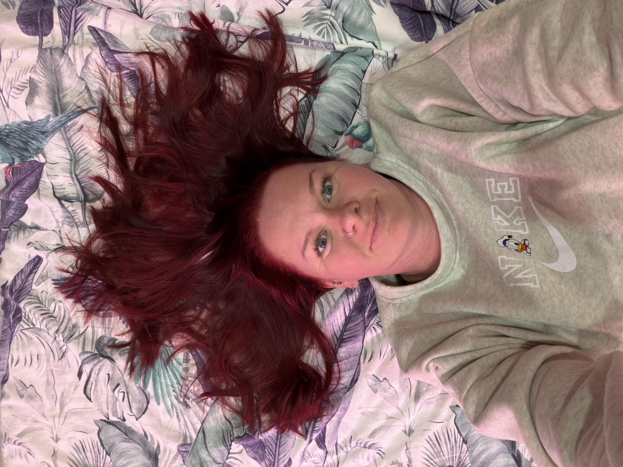A pale-skinned individual with bright blue eyes and dyed dark red hair is depicted lying on their back, looking directly at the camera with a slight smile. Their red hair is fanned out dramatically around their head on the surface below. The individual wears a light grey crew-neck sweatshirt featuring a white "NIKE" logo on the chest, with a small cartoon depiction of Donald Duck's head integrated into the 'I' of "NIKE". A silver hoop nose ring is present on the left nostril, and multiple silver earrings are visible on the left ear, including two hoops and a stud. The person is situated on bedding with a light background and a tropical or botanical pattern comprising large stylized leaves in shades of green, purple, and grey, along with partial depictions of green and blue bird-like figures. The scene is an indoor setting; the specific city of Chaikovsky, Russia, is not discernible from the visual information presented.