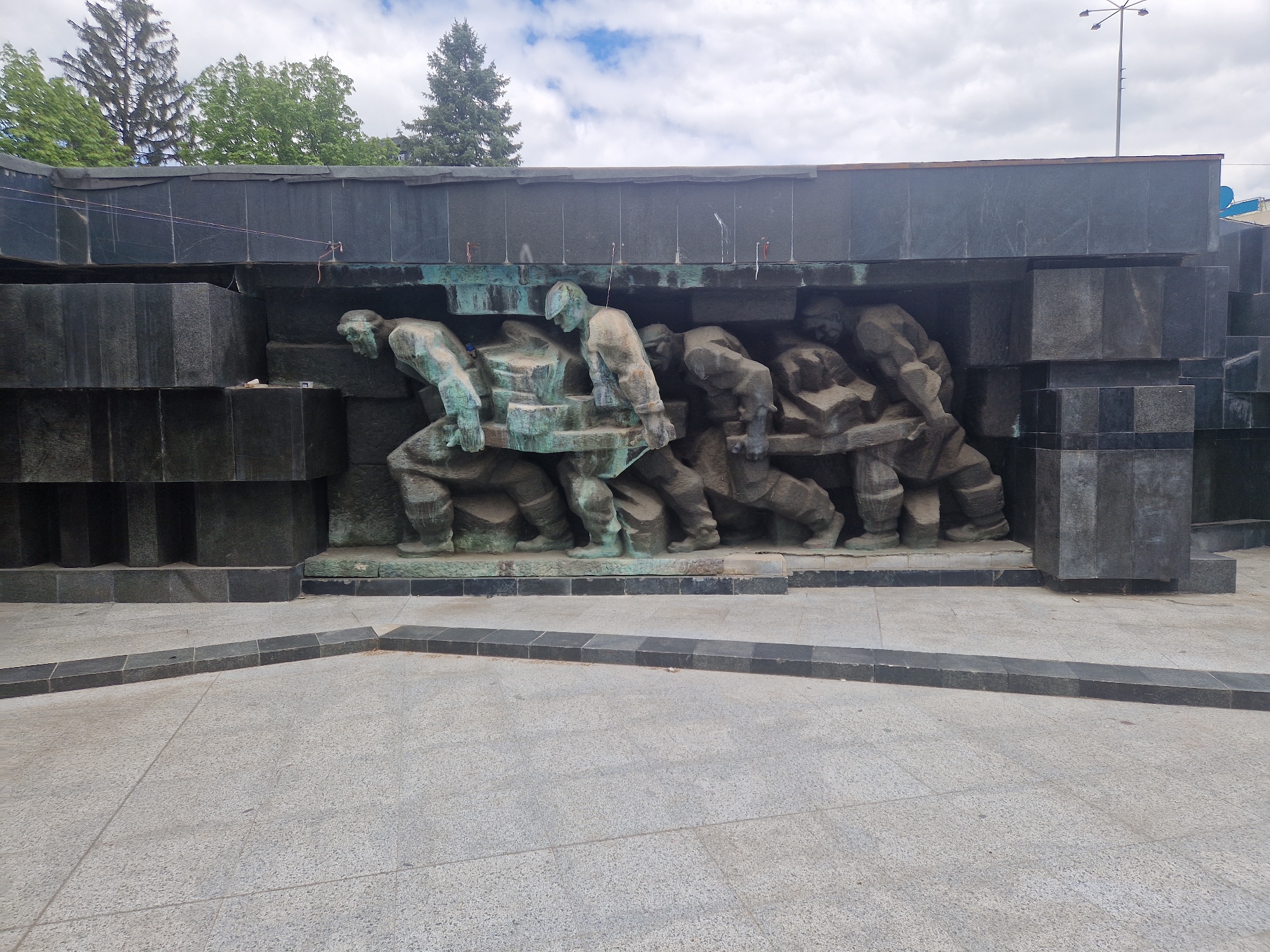 The image shows a monument in an urban area. The monument depicts a group of six figures appearing to be laborers, cast in bronze, carrying a large, undefined object. The figures are positioned under a dark, granite-like structure made of rectangular blocks. A paved area stretches in front of the monument. In the background, trees and a cloudy sky are visible. The monument appears to be in a city, but the specific location remains Unknown, Unknown.