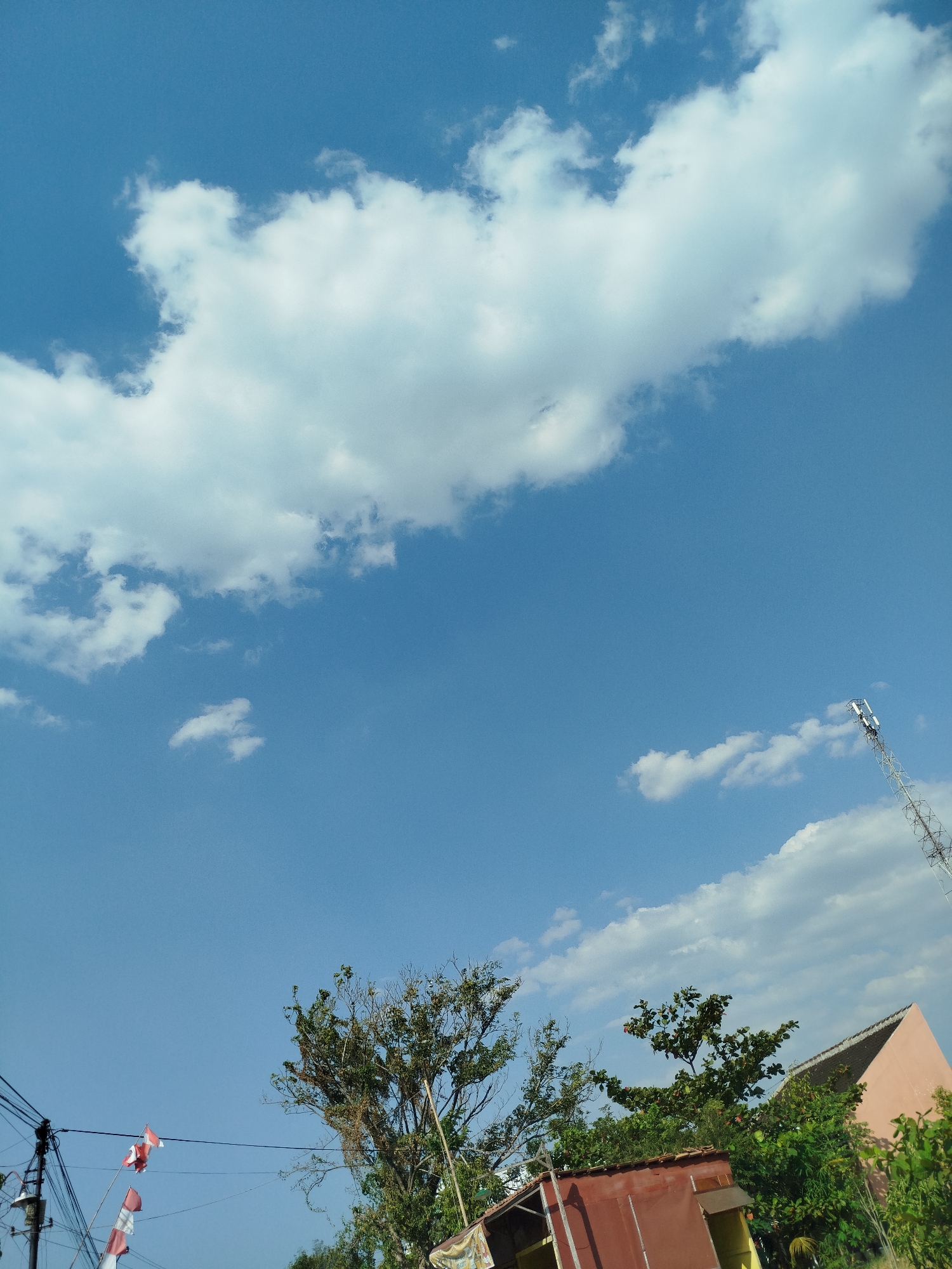 A low-angle, medium shot captures a clear blue sky with scattered white cumulus clouds over the city of Mororejo, Indonesia. In the upper right portion of the frame, a tall, metallic telecommunications tower stands against the sky, its structure a lattice of white and red. Below and to the left of the tower, the sloped roof of a light peach-colored building is visible, partially obscured by lush green foliage. In the foreground, a predominantly red, low-rise structure with a corrugated metal roof is seen, its yellow doorway visible. To the left of this structure, a utility pole with numerous electrical wires is present. Attached to these wires are several red and white Indonesian flags, fluttering in the breeze. The overall scene is one of a partly cloudy day, with a mix of urban infrastructure and natural elements.