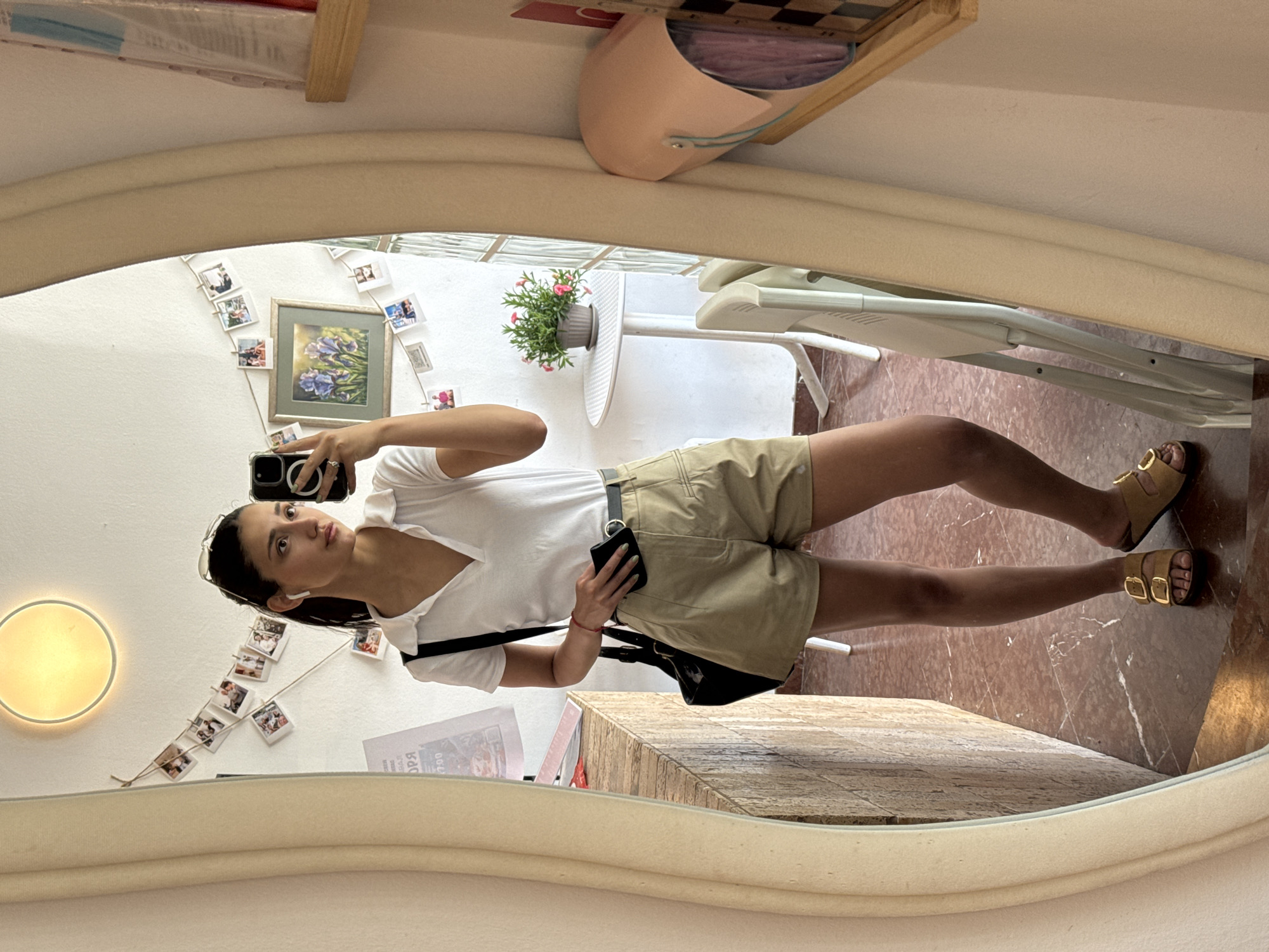 The image depicts a young woman taking a selfie in a mirror. She is wearing a white v-neck t-shirt, khaki shorts with a black belt, and tan sandals with gold buckles. She is holding a black smartphone in her left hand, and a small black purse or wallet in her right. A black shoulder bag hangs across her body.
The reflection reveals a room with white walls adorned with a string of small photographs and a framed print. A round light fixture is visible on the wall. A white table with a potted plant sits near a white high chair. The floor is covered in reddish-brown marble.
Elements suggesting the location might be the city of Bar, Montenegro, are not readily apparent within the image, but the architectural style, materials, and interior design do not contradict the possibility.