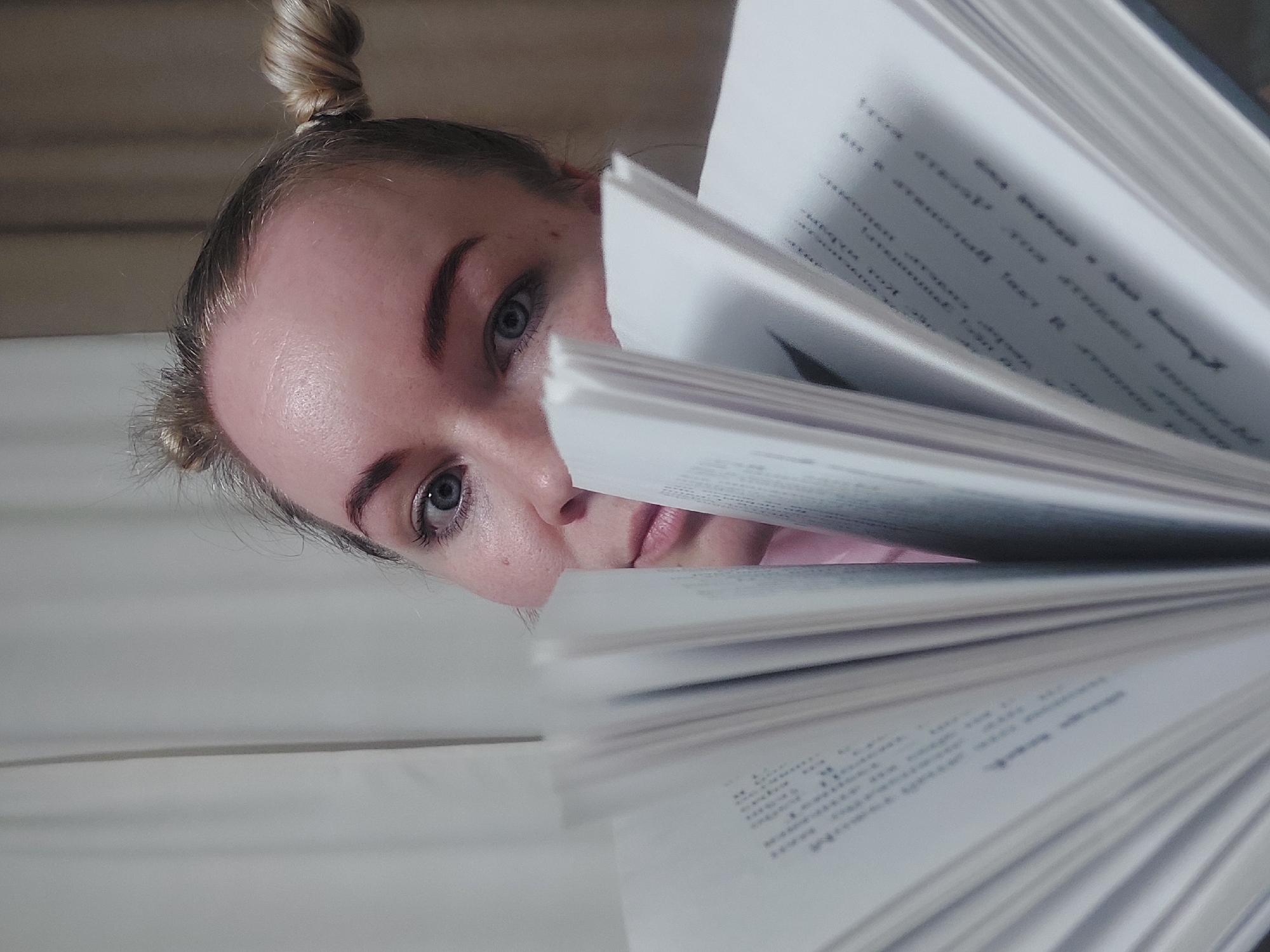 A pale-skinned woman with blonde hair styled in two small buns on top of her head is partially visible behind a large, open book. Her face is angled downwards, with her blue eyes, nose, and lips visible. The book fills a significant portion of the frame, and the text on the white pages is blurred. The background appears to be a light-colored wall or surface. The scene appears to be indoors. There is no information available to determine the location as the city of Chaikovsky, Russia.