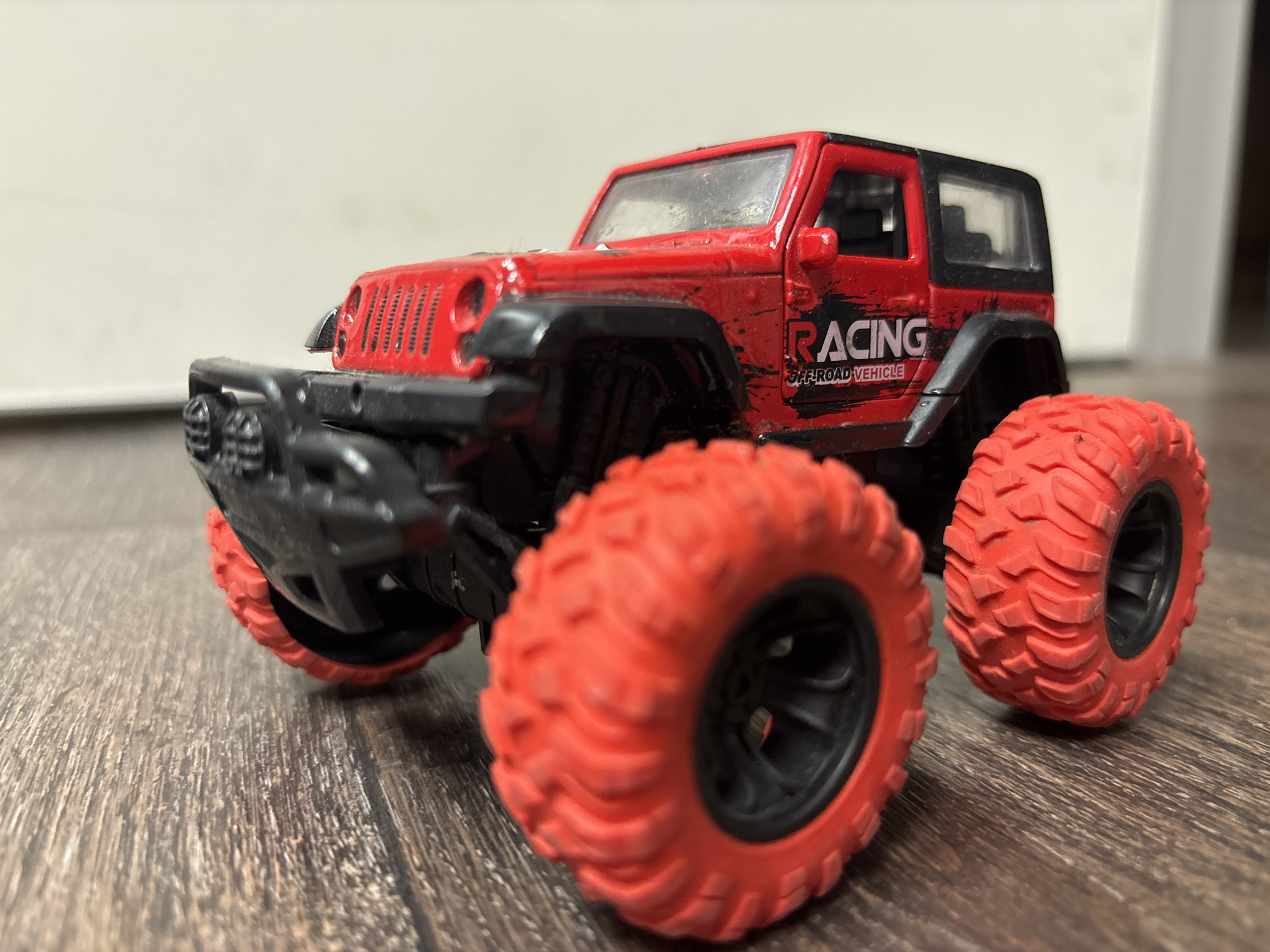 The image displays a close-up, low-angle shot of a red toy off-road vehicle, resembling a Jeep Wrangler. The vehicle is predominantly red with a black roof and black trim around the wheel wells. Prominently visible on the side of the vehicle is the word "RACING" in white block letters, with "OFF-ROAD VEHICLE" written below it in smaller, distressed font. The toy has large, aggressive red tires with black rims. The vehicle is positioned on a wooden surface, which appears to be a floor, with the background blurred, showing a light-colored wall and a hint of a doorway. There are no people or animals present. The lighting suggests an indoor setting, possibly during the day due to the brightness. The focus is sharply on the toy vehicle, highlighting its rugged design and bold coloring. The scene conveys a sense of playfulness and adventure, associated with off-road racing.