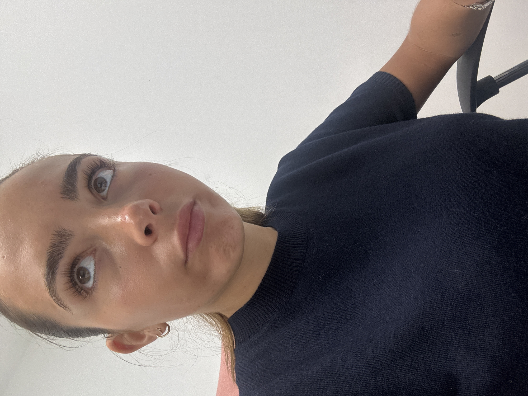 The image depicts a woman looking upward at the camera. She has fair skin, brown eyes, and dark eyebrows. She is wearing a dark blue knit top. Her hair appears to be pulled back, and she is wearing small hoop earrings. The background is a plain white wall. The location is not discernible from the provided image.