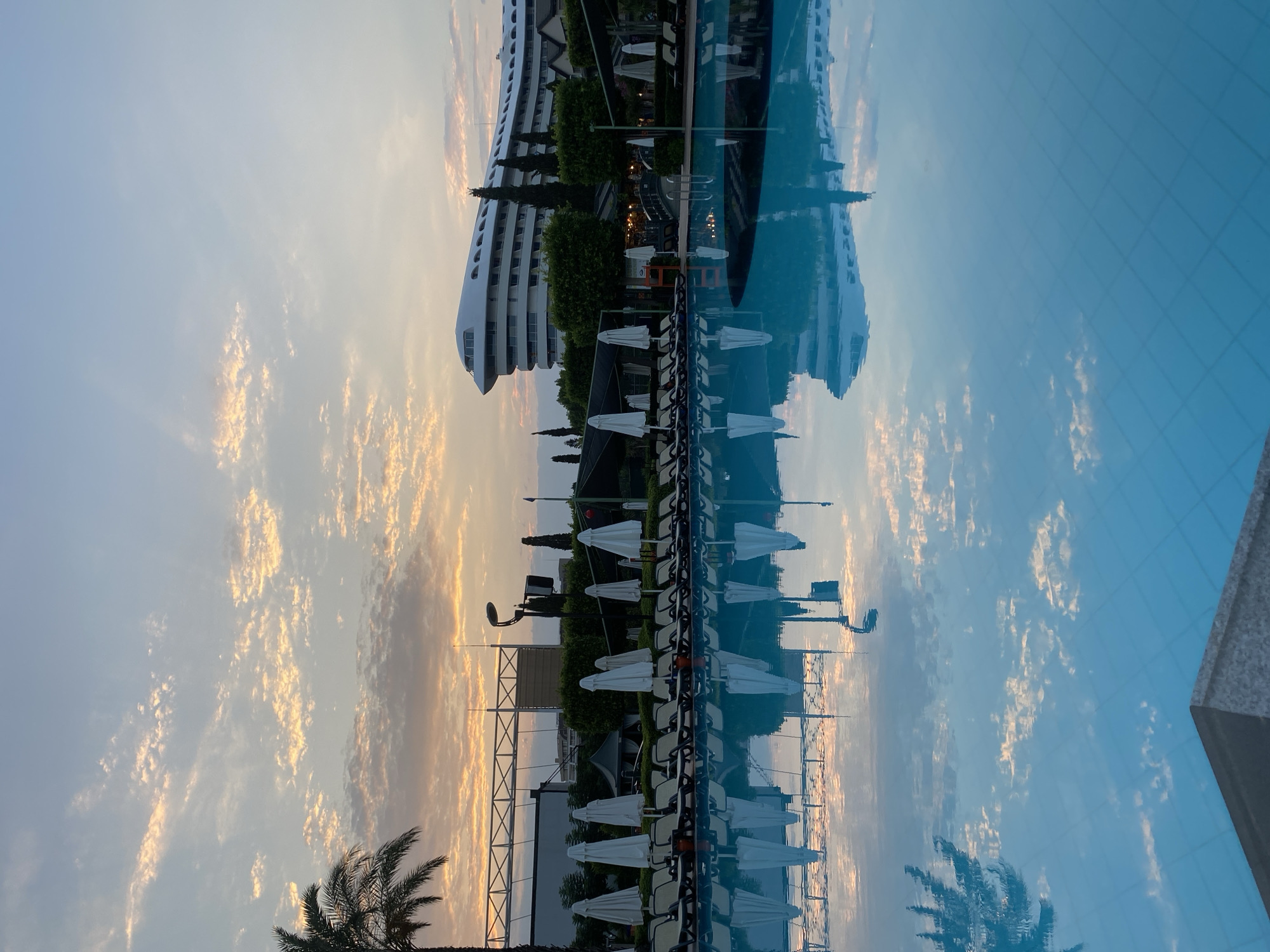 A serene shot captures an empty swimming pool at what appears to be a resort in Aksu, Turkey, during sunrise or sunset. The water, a vibrant turquoise, mirrors the soft hues of the sky, which is painted with wispy clouds tinged with orange and pink. Rows of closed white umbrellas and lounge chairs line the pool's edge, suggesting a calm morning or evening. In the background, a large, modern hotel with a distinctive curved white facade stands tall, surrounded by lush green trees and palm trees. A distant stadium structure is also visible, hinting at a lively location. The overall ambiance is tranquil and inviting, showcasing the beauty of a resort setting in Aksu, Turkey.