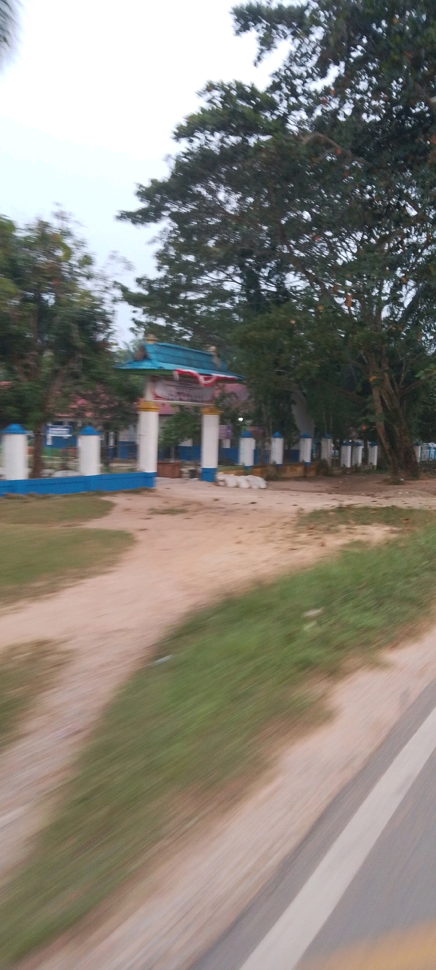 The image depicts an entryway with a blue and white striped gate and a tiled blue roof. A sign with Indonesian writing is visible above the entrance, adorned with red and white Indonesian flags. The entryway is flanked by tall white pillars with blue accents. Beyond the gate, a building with a blue facade and white windows is visible, suggesting it is a school or public institution. The ground is a mixture of dirt and grass. Trees, some with dense foliage, surround the area. In the foreground, a blurred dirt road with a grassy edge is visible, indicating movement or a slight motion blur in the photograph. The overall setting appears to be in Lenggadai Hulu, Indonesia, likely a public building entrance.