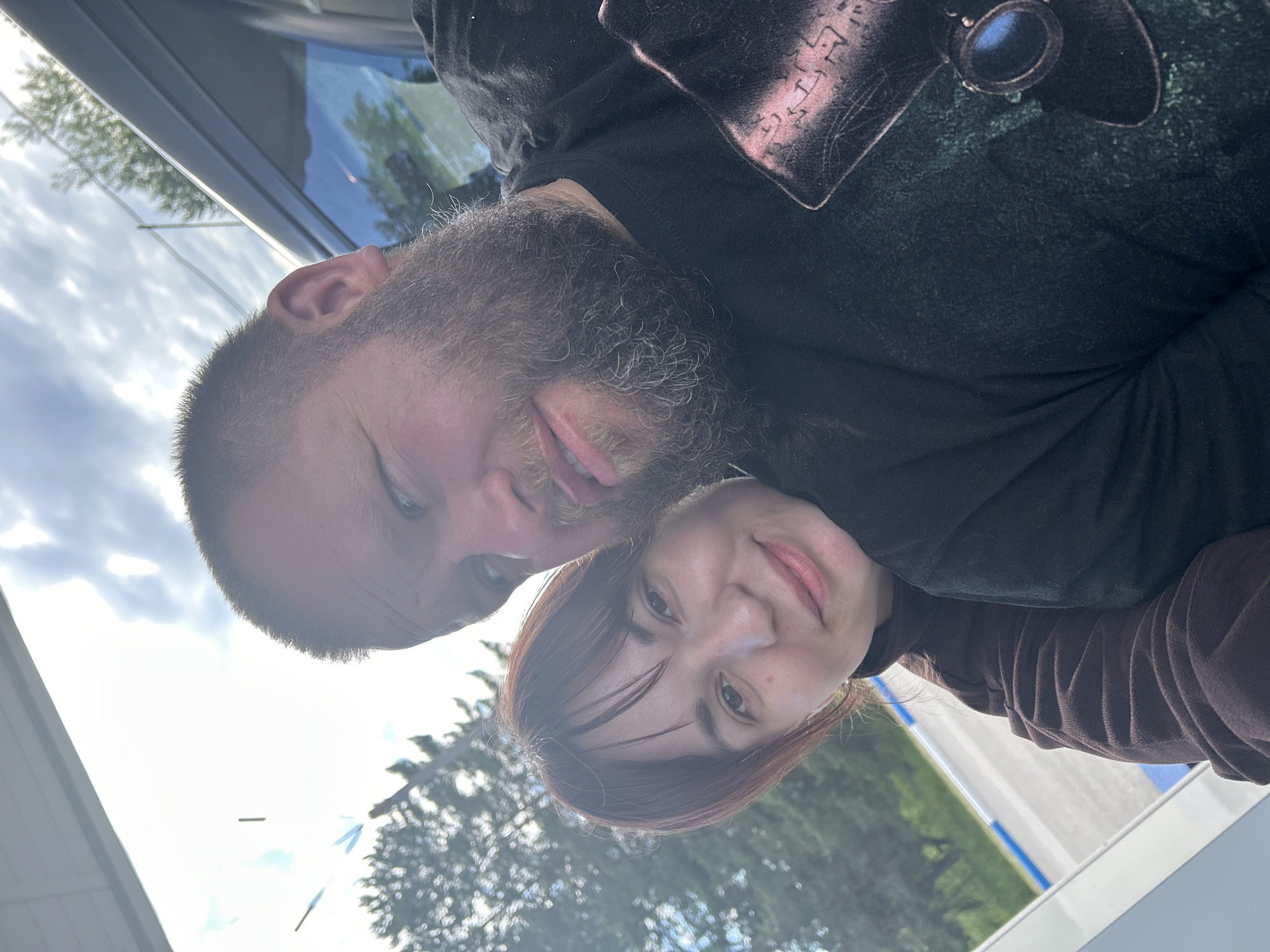 The image is a rotated close-up selfie featuring two individuals, a man and a woman, set against a bright, hazy sky and blurred outdoor elements. The picture is oriented such that the subjects' heads are tilted towards the right, suggesting it was originally taken in portrait mode and then rotated 90 degrees counter-clockwise.
On the left, a man with a prominent, dark, and graying beard is visible from the forehead down. He has dark eyes and a slight, open-mouthed smile, showing a hint of his tongue. He is wearing a dark-colored shirt that features a faded or bleached pattern on the shoulder and chest, displaying abstract, stylized symbols or glyphs. A pair of dark-lensed sunglasses is partially visible, seemingly hanging from his shirt near the top right of the frame.
Next to him on the right, a woman with shoulder-length reddish-brown hair and bangs frames her face. She has dark eyes and a gentle, closed-lip smile. She appears to be wearing a dark top, possibly a turtleneck, layered under a brown outer garment with noticeable vertical folds or pleats on the sleeve.
The background consists of a bright, somewhat overcast sky, with reflections of green trees visible in a glass surface, likely a vehicle window or a building facade. The strong, diffused lighting indicates it is daytime. Both individuals convey a sense of contentment and relaxation. The setting implies an outdoor location or within a vehicle, situated in Chaikovsky, Russia.