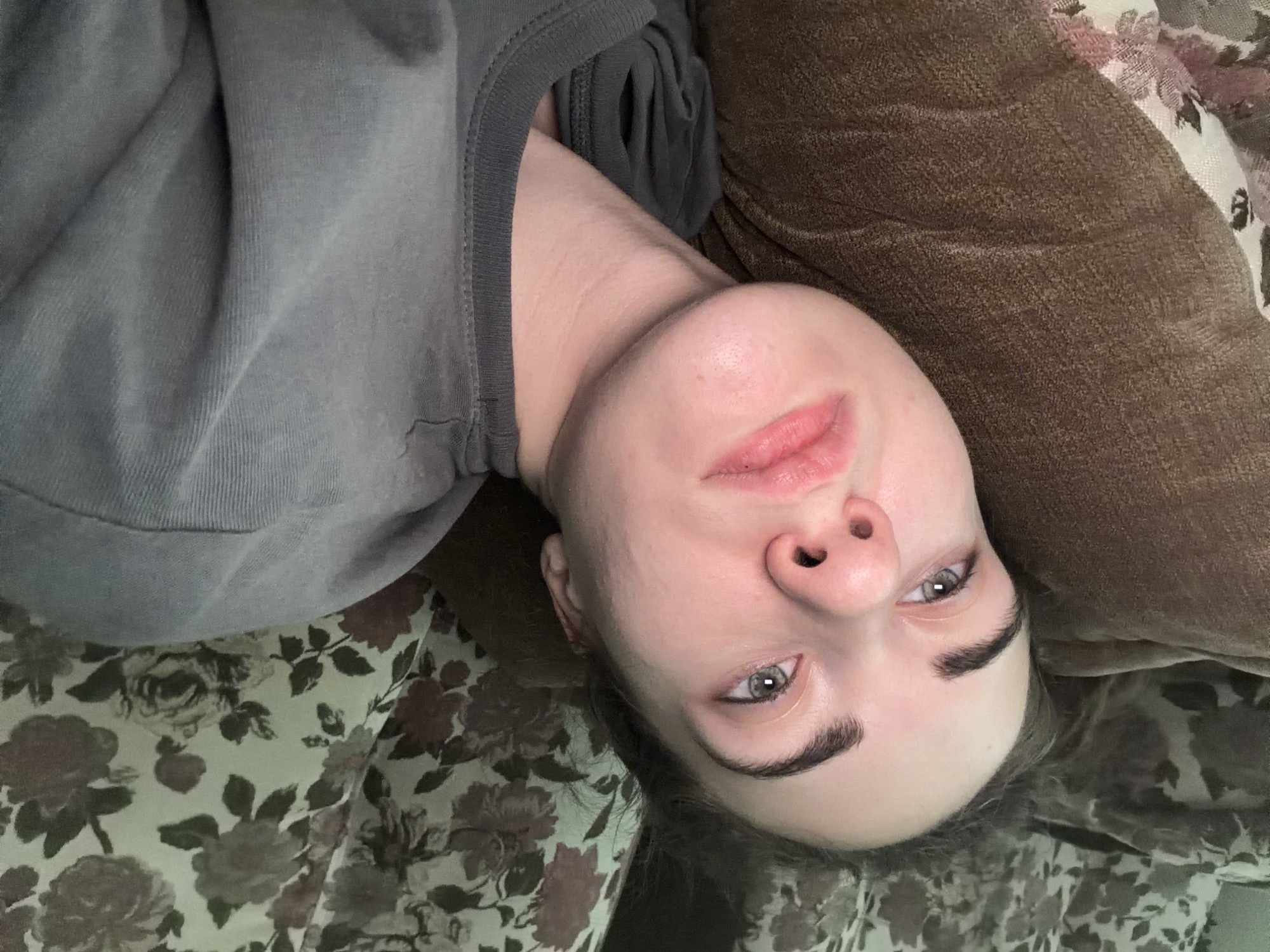 The image shows a close-up, high-angle shot of a young woman lying down. She is resting her head on a brown textured surface, possibly a pillow or cushion. Behind her, a floral patterned fabric, likely part of bedding or upholstery, is visible. The woman is wearing a gray t-shirt. Her expression is neutral, and her eyes are open, looking upwards. The lighting appears to be natural, possibly from a window. No specific details about Zavodoukovsk, Russia, or any city-related elements are present in the image.