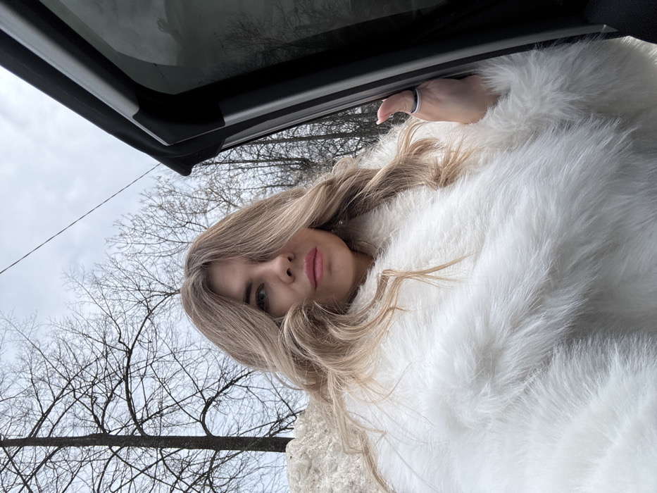 A fair-skinned female, with long, wavy, light brown or blonde hair obscuring the right side of her face, is depicted in a close-up, chest-up shot. She wears pink lipstick and a voluminous white faux fur coat. Her left hand, adorned with a narrow bicolor ring (dark and light bands) on the index finger, grips the vertical edge of an open, dark-colored vehicle door, indicating interaction with a car. The background consists of bare deciduous trees and a prominent pile of melting snow on the left, under an overcast sky. The environment suggests a late winter or early spring outdoor scene. The setting is located in the city of Chaikovsky, Russia.