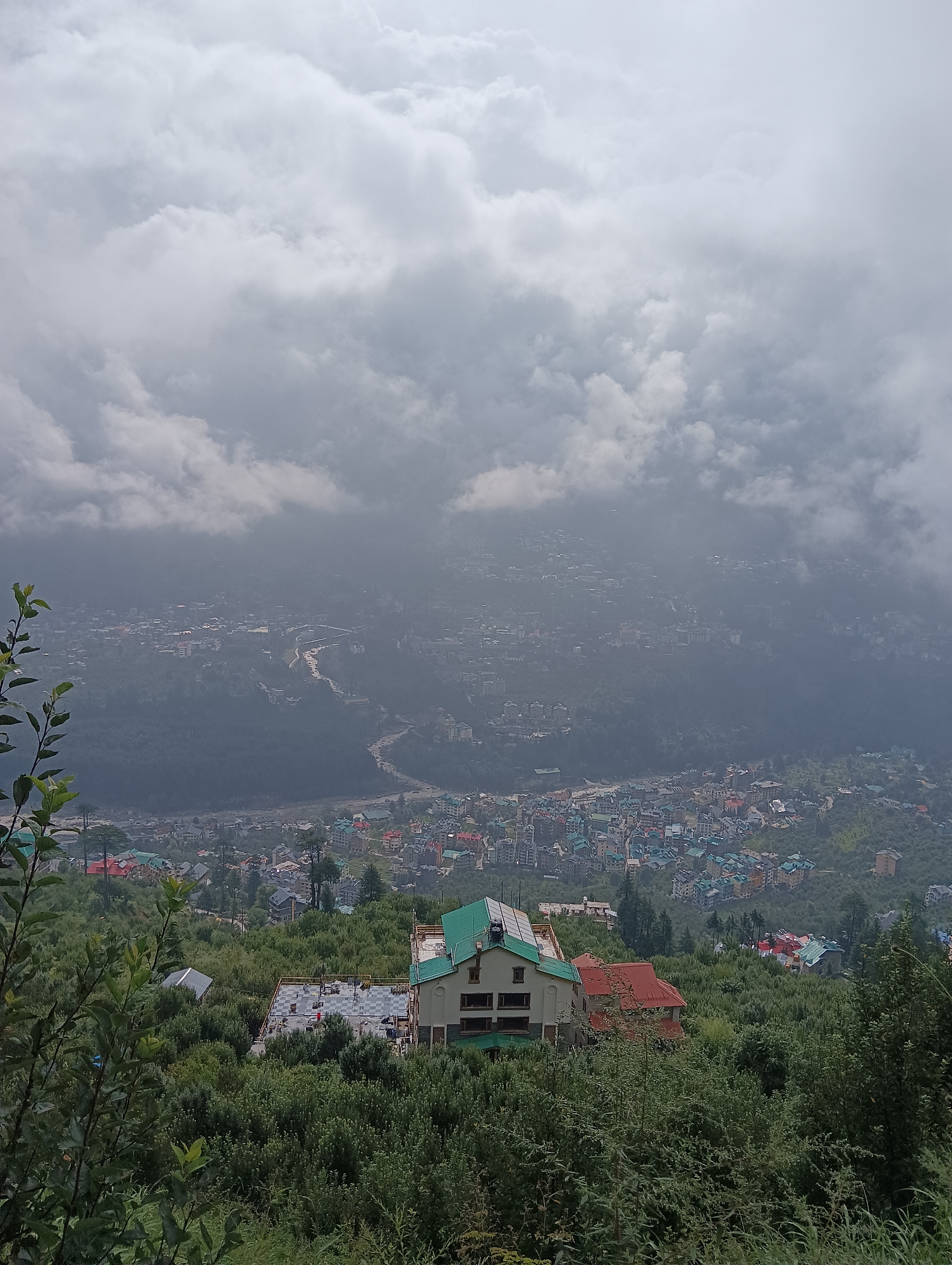 The image shows a panoramic view of the city of Siumsa, India, nestled in a valley. The foreground is dominated by lush green foliage and trees, with a partially constructed building visible on the right. This building has a light-colored facade, a dark-tiled roof, and solar panels installed on its upper section. In the middle ground, a cluster of houses with colorful roofs, predominantly red and green, are scattered amidst the dense greenery. The background reveals the sprawling city of Siumsa, characterized by a dense arrangement of buildings with varied architectural styles and roof colors. A river or road winds through the cityscape, disappearing into the hazy distance. The upper portion of the image is filled with a dramatic sky, featuring thick, white and gray clouds that partially obscure the view of the distant mountains and the sky itself. The overall atmosphere is one of a serene, mountainous landscape enveloped in a slightly overcast or foggy condition. No people are visible in the image.