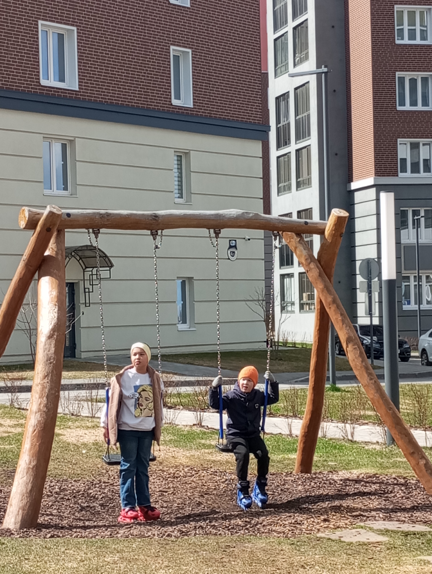 Two children are at a playground in Pervomaysky, Russia. One child stands on a swing, while the other is seated and swinging. The playground is located in a residential area. Behind the playground are several buildings. Cars are parked on the street beyond the playground.
