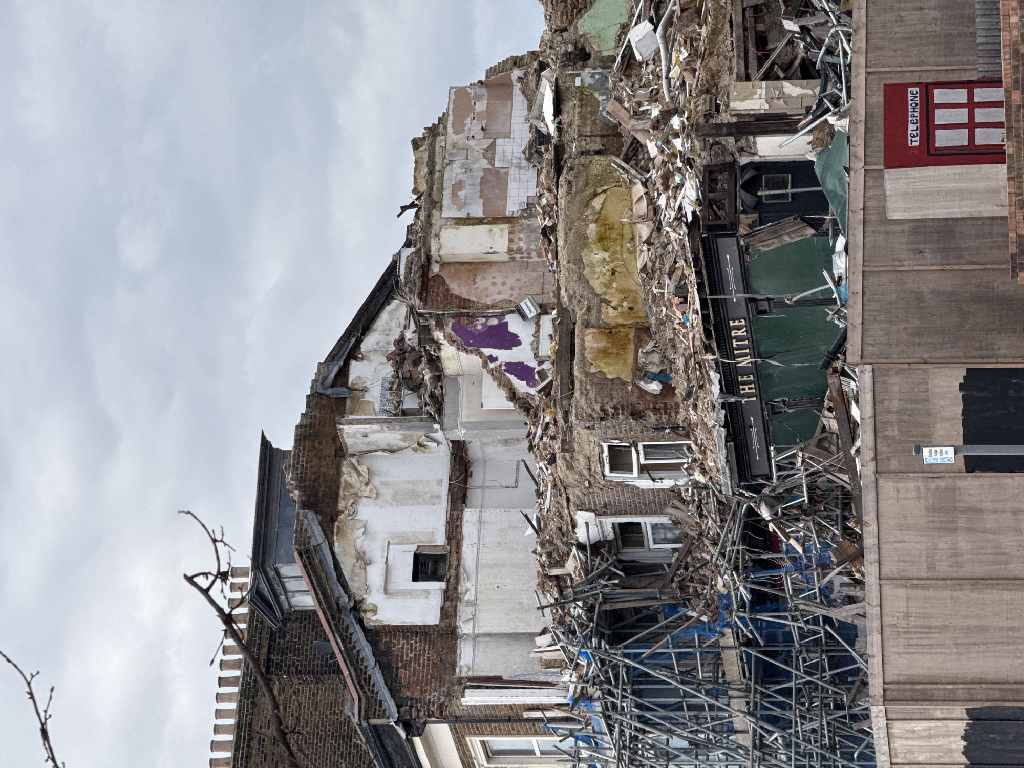 The image depicts a building in partial demolition. Signage indicates "THE MITRE". A painted red telephone box is visible on a adjacent wall. Scaffolding and debris surround the structure. Location suggested is London, United Kingdom. No people are apparent.