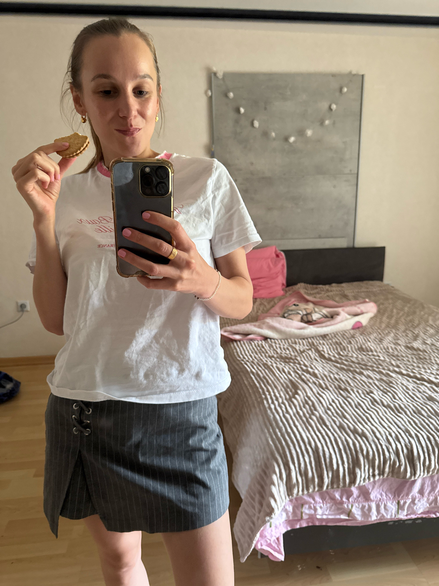 A young woman, with blonde hair pulled back, stands in a bedroom and holds a partially eaten cookie in her left hand, which is raised to her mouth. She is wearing a white t-shirt with pink trim and a gray pinstripe skirt with lace-up detailing. Her right hand holds a dark smartphone, displaying its screen to the viewer. The background shows a bed with a patterned comforter and a pink pillow. A grey headboard is visible behind the bed, and a concrete-like wall hangs behind the woman, decorated with what appears to be string lights. The overall lighting suggests it is daytime. The setting is a bedroom, likely in a residential apartment or house. No specific landmarks or indicators of the city of Überherrn, Germany are present in the image.