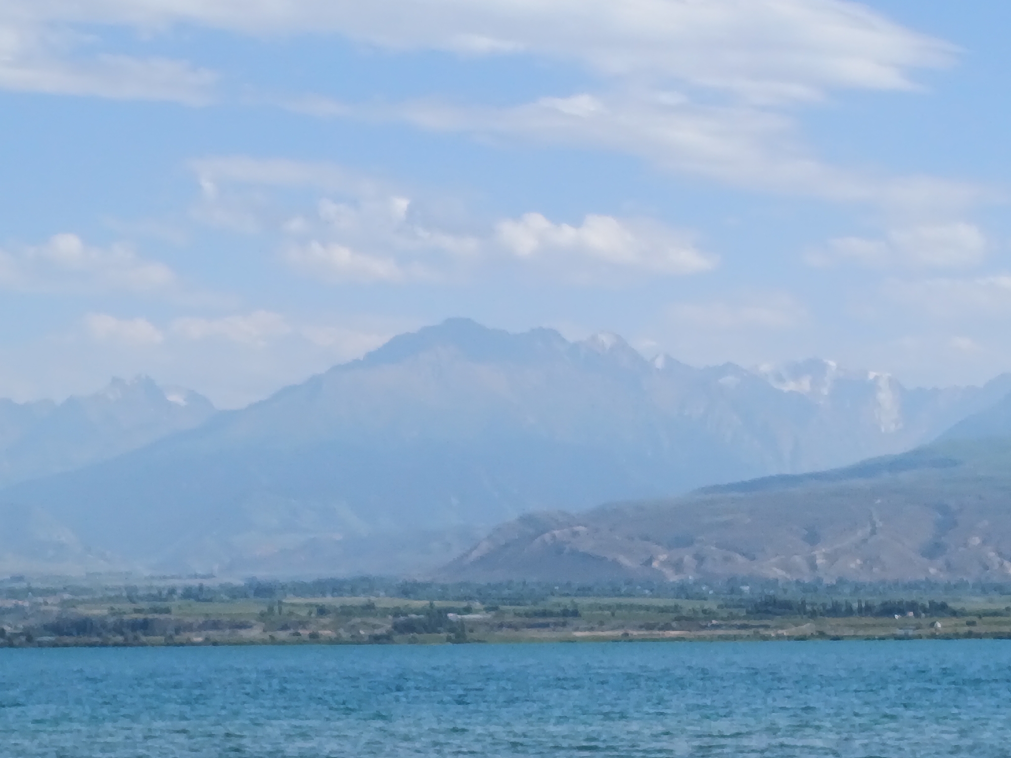 This image depicts a tranquil natural landscape. In the foreground, a body of water with a turquoise hue stretches across the frame, its surface gently rippled. Beyond the water, a verdant shoreline is visible, dotted with scattered trees and greenery. Further in the distance, a range of mountains rises, their peaks softened by atmospheric haze. The sky above is a pale blue, adorned with scattered white clouds. The overall scene conveys a sense of peacefulness and natural beauty. The city of Kirpichny, Kyrgyzstan is known for its mountainous terrain and proximity to water bodies like Issyk-Kul Lake, which this image could potentially represent. However, without further context or specific landmarks, it is impossible to definitively confirm the location.