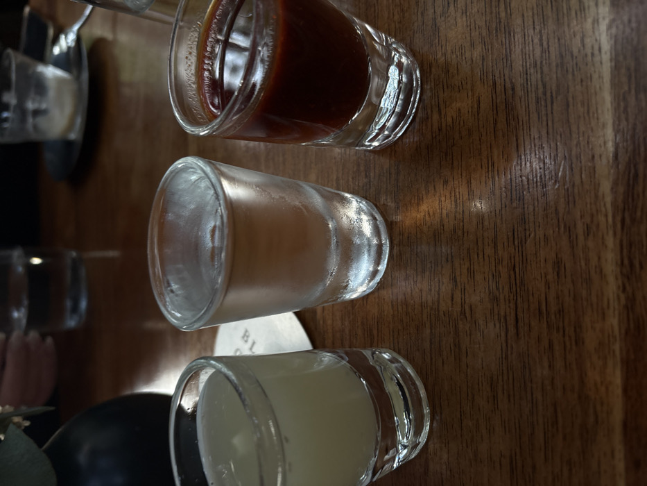 Three clear shot glasses line a wooden table. The closest glass contains a white opaque liquid. The second glass has a tan liquid, with condensation covering the glass. The farthest glass holds a dark brown liquid. Two more shot glasses are visible in the background, one containing a clear liquid, the other an opaque white substance. A black sphere and green foliage are visible beside the shot glass containing white liquid.