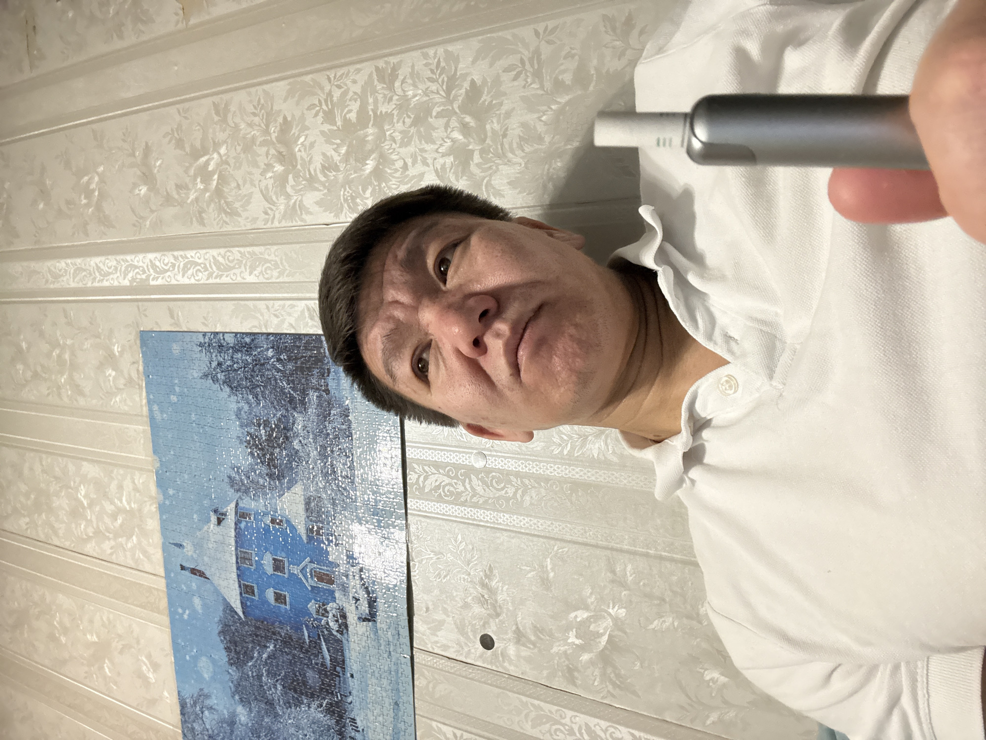 A man wearing a white collared shirt is holding a vaping device. A puzzle with a winter scene hangs on the wall. The wallpaper has a floral design. Based on prior knowledge, this image was captured in Khimki, Russia.