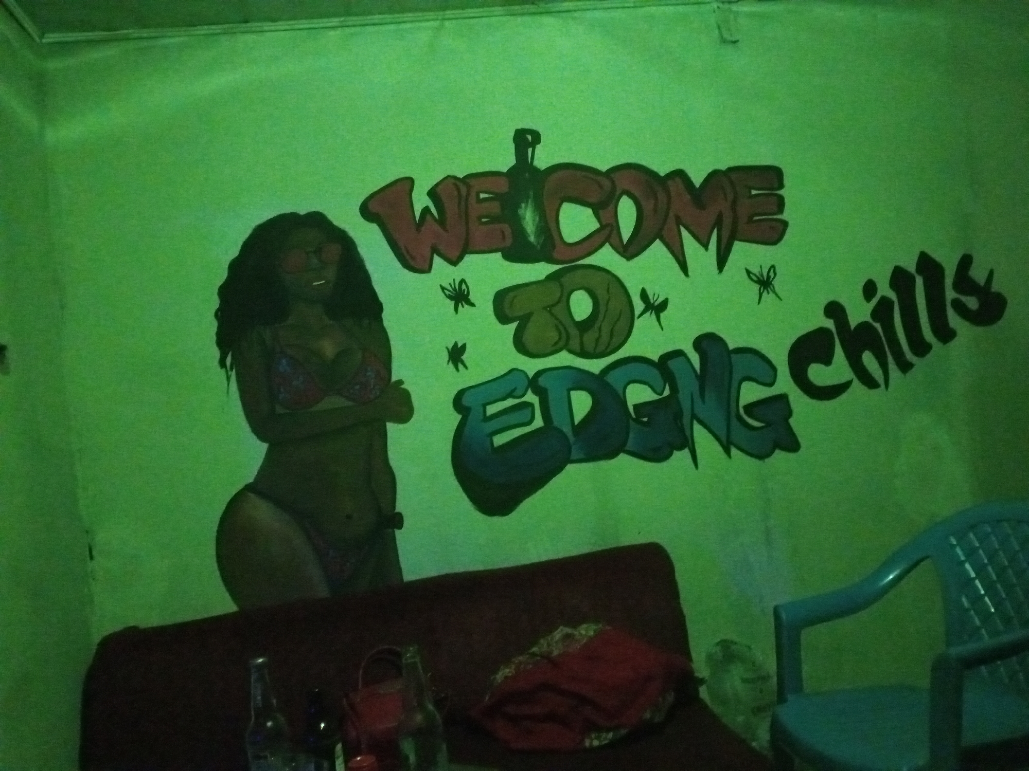 The image depicts the interior of a room with a green-painted wall displaying graffiti art. The graffiti includes the words "Welcome to Edging Chills," with an image of a dark-skinned woman in a bikini painted on the left. Below the graffiti art are a red couch, bottles, a bag, and a blue plastic chair. The location is in Jos, Nigeria.