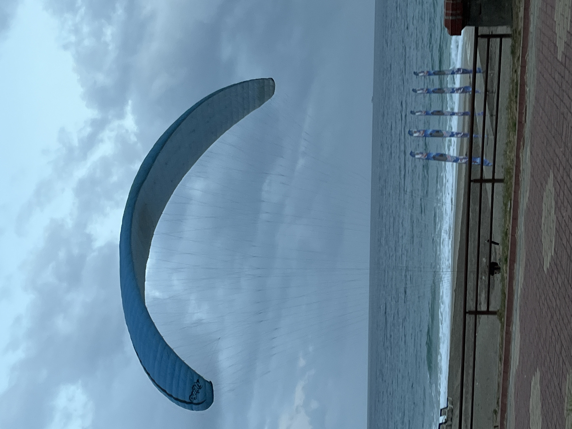 The image depicts a beach scene in Alanya, Turkey. A blue and white kitesurfing kite is prominently displayed against a cloudy sky, partially obscuring the view of the water and shore. In the background, the sea is visible, with waves breaking on the sandy beach. A low wall runs along the edge of the beach, with black metal railings. Five cylindrical banners are attached to the railings. A section of red brick pathway borders the wall.