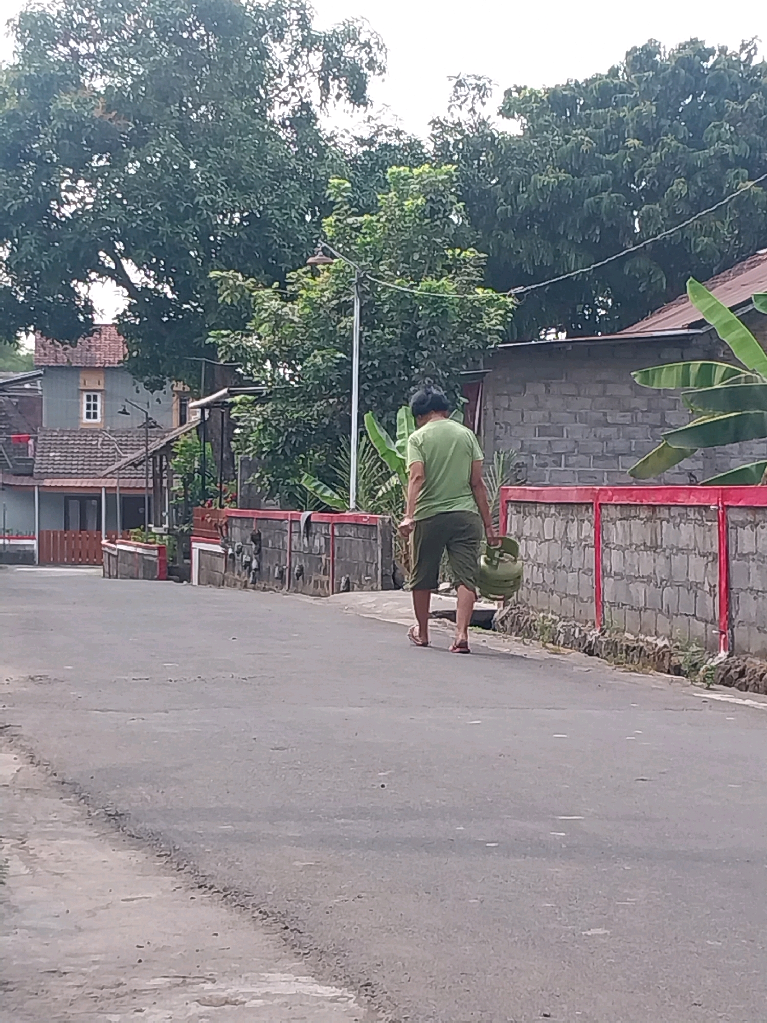 The image shows a person walking down a paved road in Purwomartani, Indonesia. The person is wearing a green t-shirt and green shorts and is carrying a green gas cylinder. They are walking away from the camera, towards a brick wall with red trim. There are houses and trees in the background. The road is asphalt and appears to be in good condition. The weather is overcast.