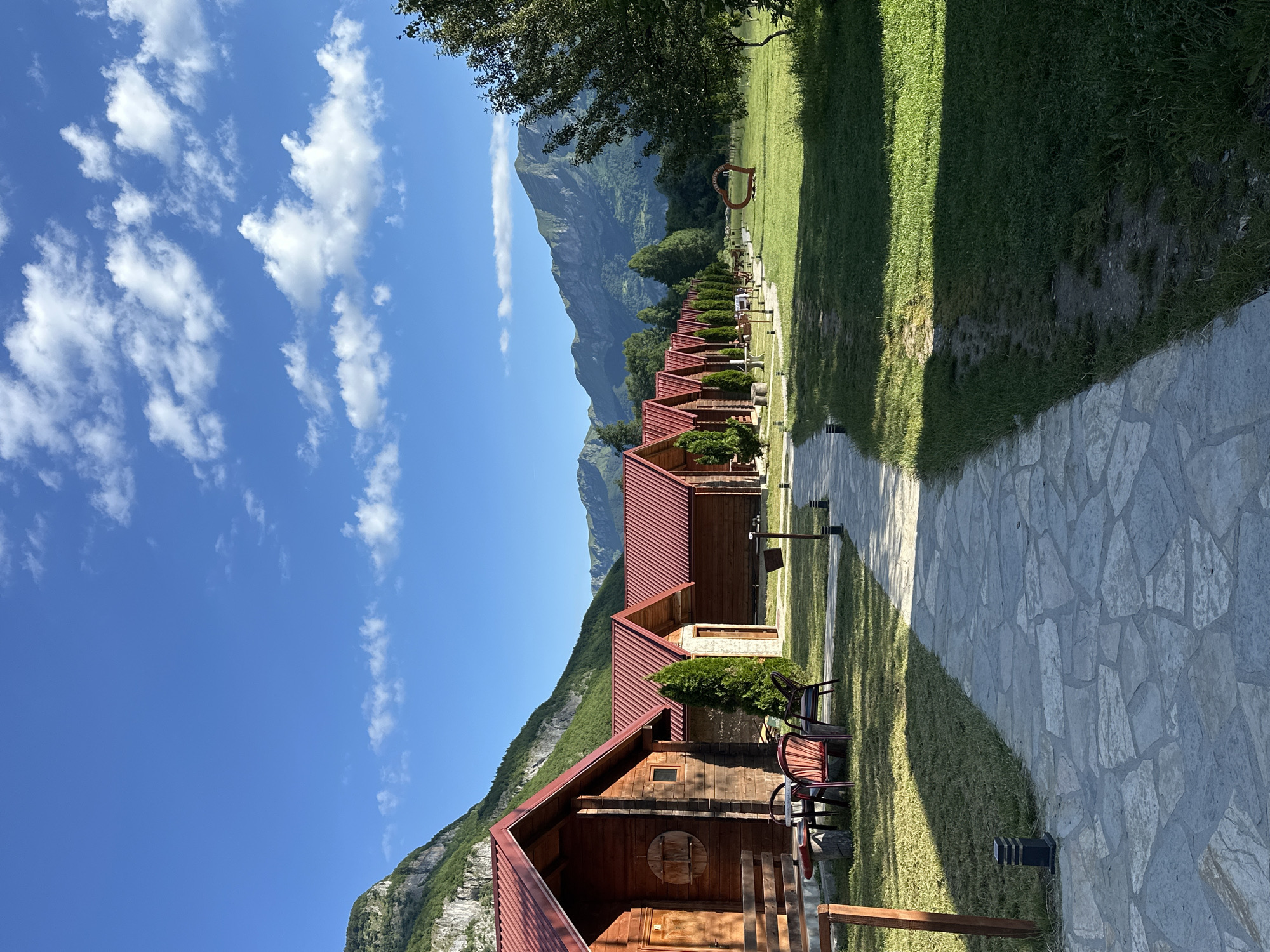 The image depicts a row of identical wooden cabins with red roofs, situated in the city of Gusinje, Montenegro. A stone pathway leads through a manicured lawn, bordered by a line of evergreen trees, towards the cabins. In the background, lush green mountains rise under a clear blue sky dotted with white clouds. To the right of the pathway, a large, red heart-shaped sculpture is visible in the grassy field. The scene suggests a serene and picturesque setting, likely a resort or a tourist accommodation.