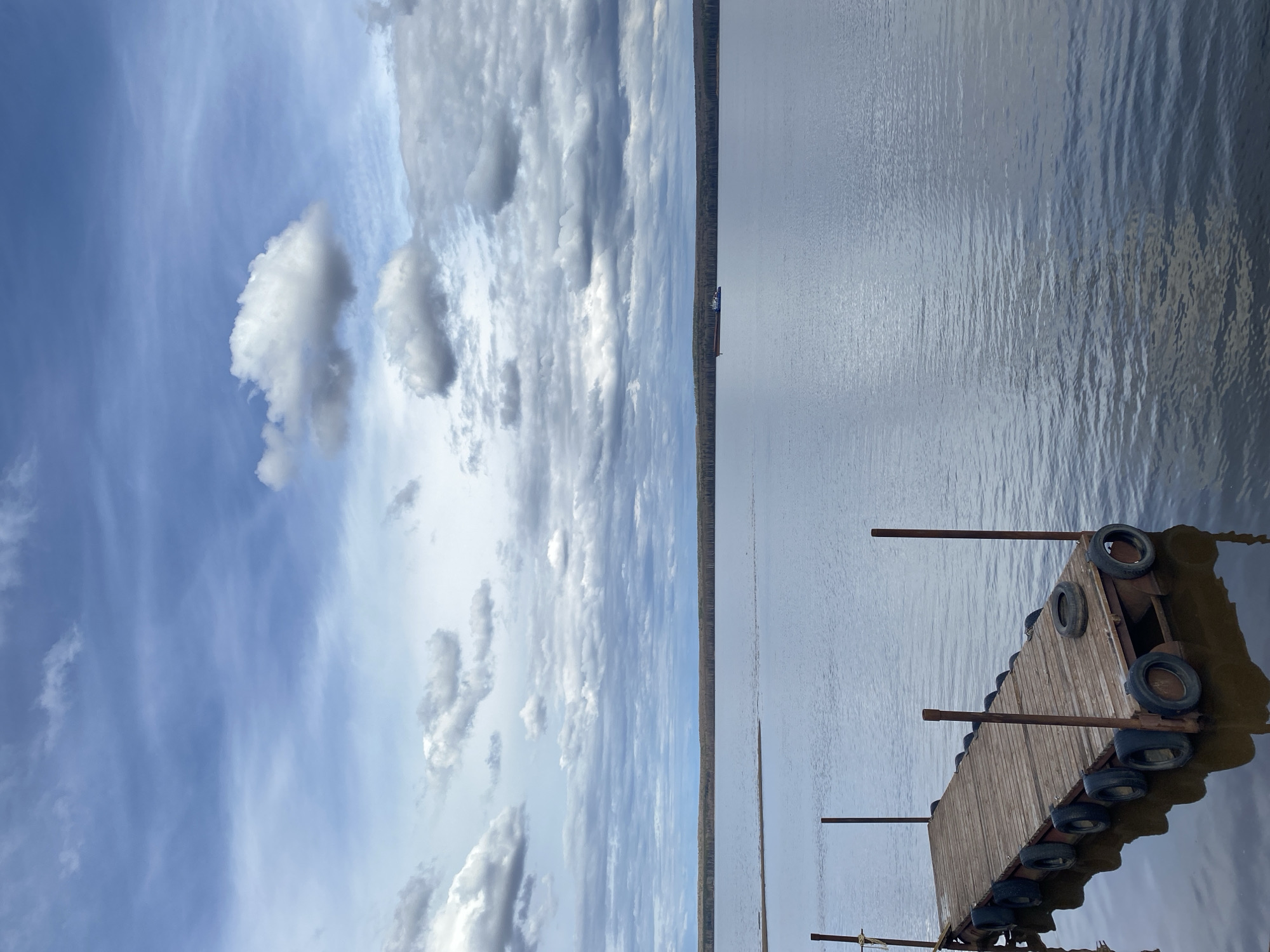 The image depicts a cloudy sky and calm body of water, likely a lake or river, with a narrow strip of land visible in the distance. In the foreground, a wooden dock or platform is visible, supported by tires and metal poles. The photograph seems to have been taken near the city of Chaikovsky, Russia, based on the likely geographical features and objects found in the area.
