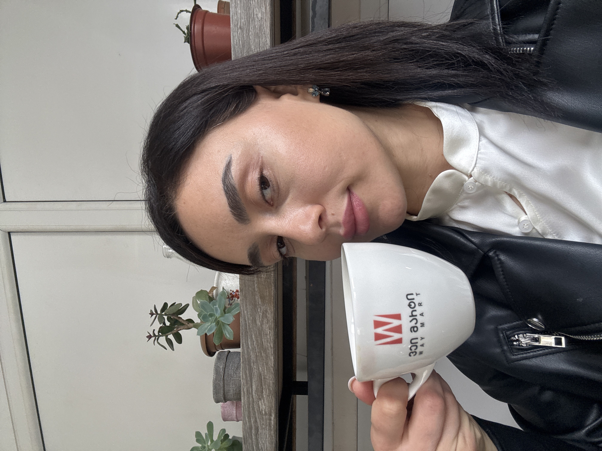 A woman with dark hair is holding a white mug with the logo of "WAY MART" and the Georgian text "ვეი მარტი" in front of a window with a shelf containing various plants in pots. She is wearing a white collared shirt under a black leather jacket. Based on the logo on the mug, this image likely was taken in Tbilisi, Georgia, as Way Mart is a supermarket chain present in the city.