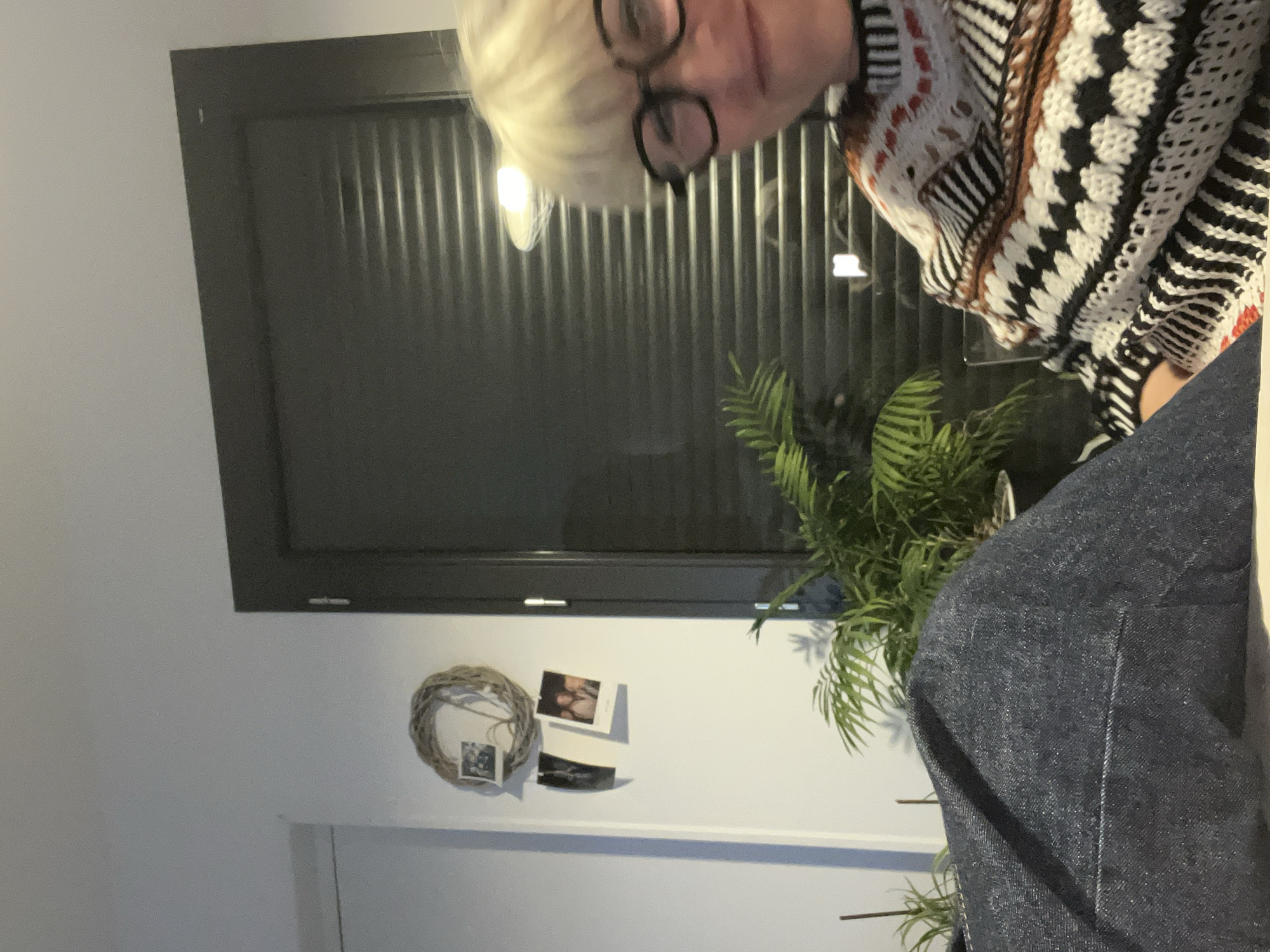 The image depicts the interior of a room. The focal point is a person with blonde hair and glasses, wearing a sweater with a black, white, and brown pattern. In the background is a dark gray window with horizontal blinds. A green plant is visible in front of the window. A wreath and several small framed pictures are mounted on the white wall beneath the window. The bottom right of the image shows a dark blue denim fabric. There is no information that can be used to confirm the location is Chambœuf, France.