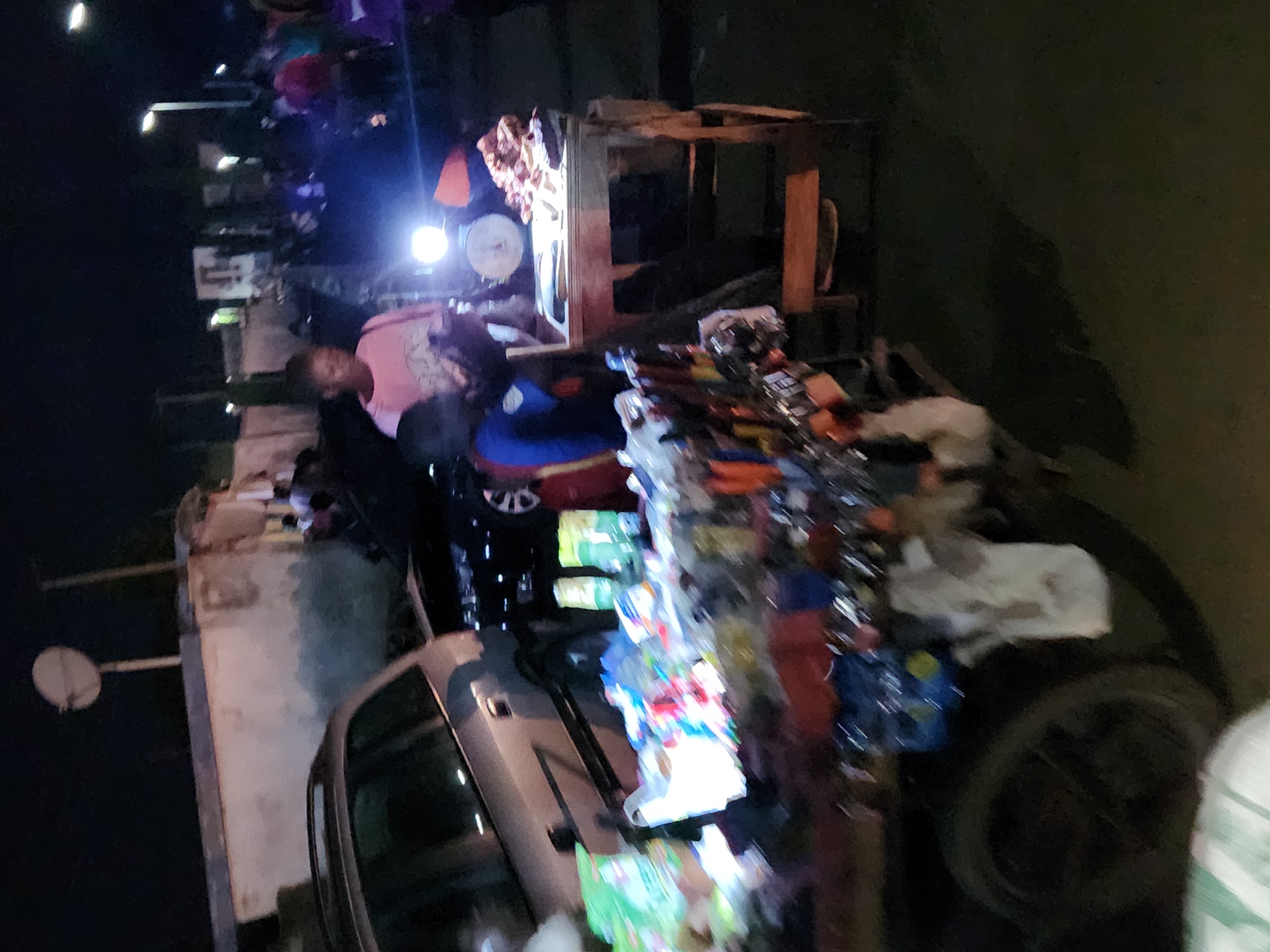 At night, a scene unfolds in the city of Kabong, Nigeria, with artificial lighting illuminating the area. In the foreground, a cart laden with various goods, including packaged items, bags, and what appears to be tools or merchandise, is prominently displayed. A wheel of the cart is visible on the left. Behind the cart, a stall with a wooden structure is set up. On this stall, a weighing scale and a pile of meat are visible. A person, likely a vendor, is standing behind the stall. To the left of the stall, a young person in a blue shirt is also present, facing the stall. Further to the left, a portion of a car is visible. In the background, more individuals are gathered, and buildings and streetlights can be seen, indicating a populated urban environment. The overall impression is that of a bustling night market or roadside commerce.