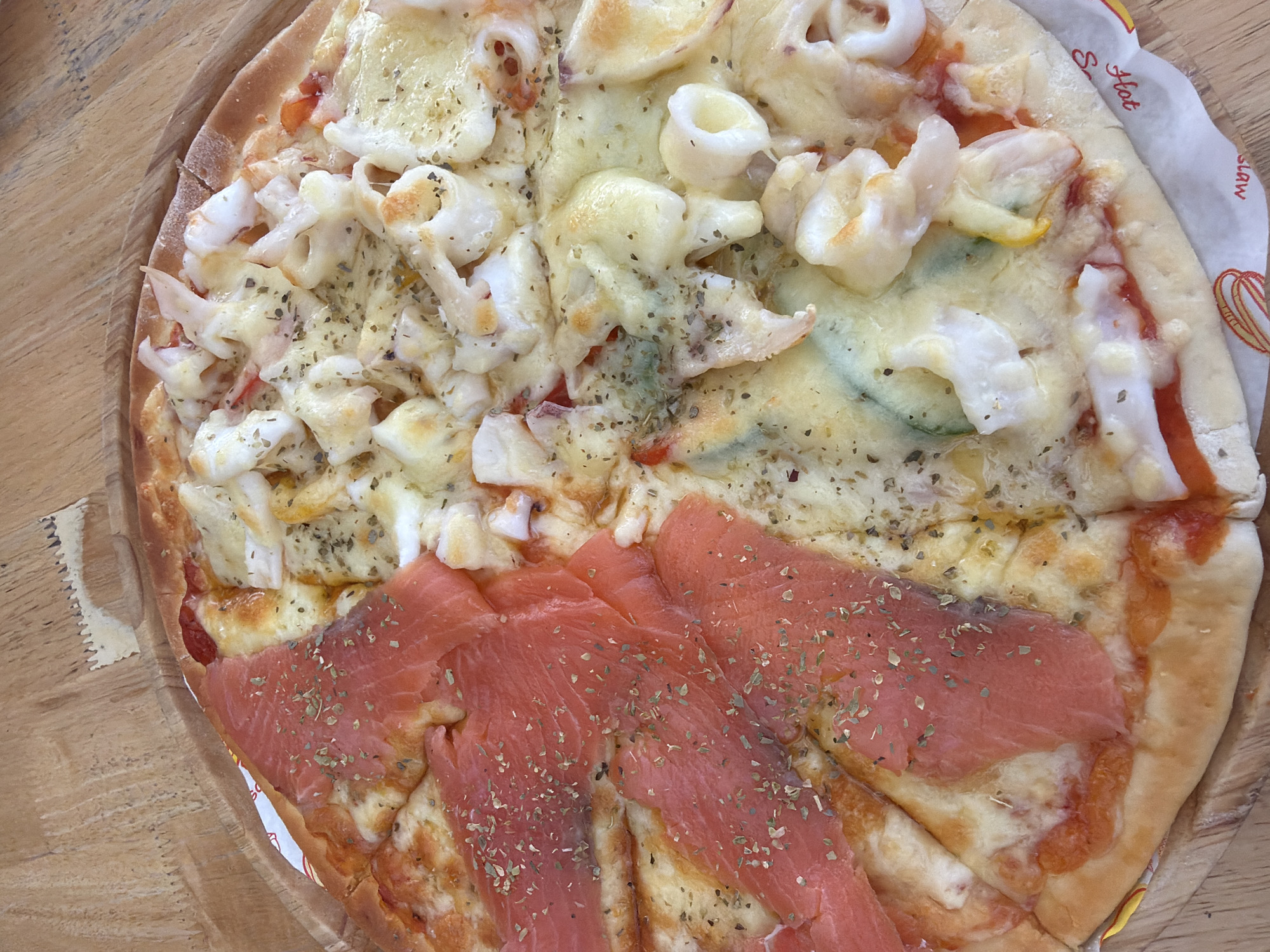 A close-up, overhead shot shows a circular pizza divided into two halves, placed on a wooden table. The left half of the pizza is topped with thin, pink slices of smoked salmon and sprinkled with dried herbs. The right half features a seafood medley including squid rings, shrimp, and possibly crab meat, covered in melted cheese and also sprinkled with herbs. The crust is golden brown and appears slightly puffy. A white paper liner with red lettering, possibly from a restaurant, is visible underneath the pizza, peeking out from the bottom edge. The background is a textured wooden surface. The scene is indoors, with natural light illuminating the pizza. The location is the Ko Samui District in Thailand. There are no people or animals present in the image.