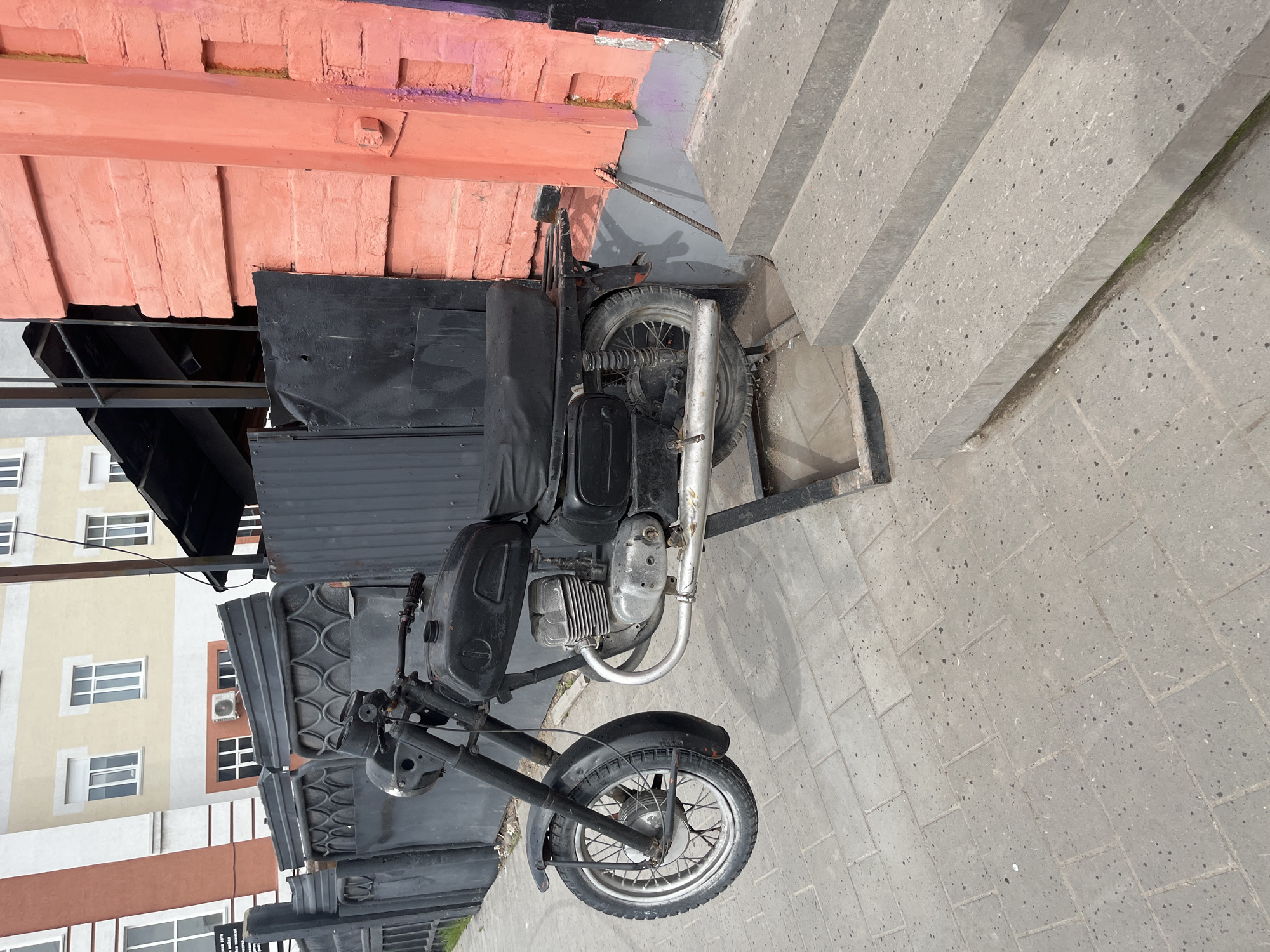 The image captures a black motorcycle parked on a paved surface next to concrete steps and a building in Ulyanovsk, Russia. The motorcycle is an older model with a visible engine, fuel tank, exhaust pipe, and spoked wheels. It is positioned against the backdrop of a salmon-colored brick wall and a gray set of concrete stairs. In the background, a multi-story building with multiple windows is visible. The setting appears to be an urban environment. There is no specific information about people in this image.