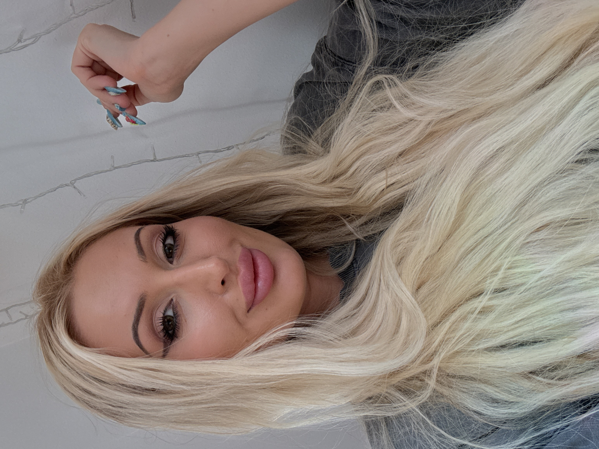 A medium shot photo shows a woman from the chest up, looking at the camera. She has long, blonde hair that is slightly wavy and reaches past her shoulders. Her eyes are dark brown, and she is wearing dark eyeliner and mascara. Her lips are plump and painted with a light pink lipstick. She is wearing a dark gray t-shirt. Her right hand is raised, and her fingers are adorned with colorful, long fingernails, with one finger touching her temple. The background is a plain white wall with faint strings of fairy lights hanging vertically. The lighting is soft, creating a warm glow on the subject.