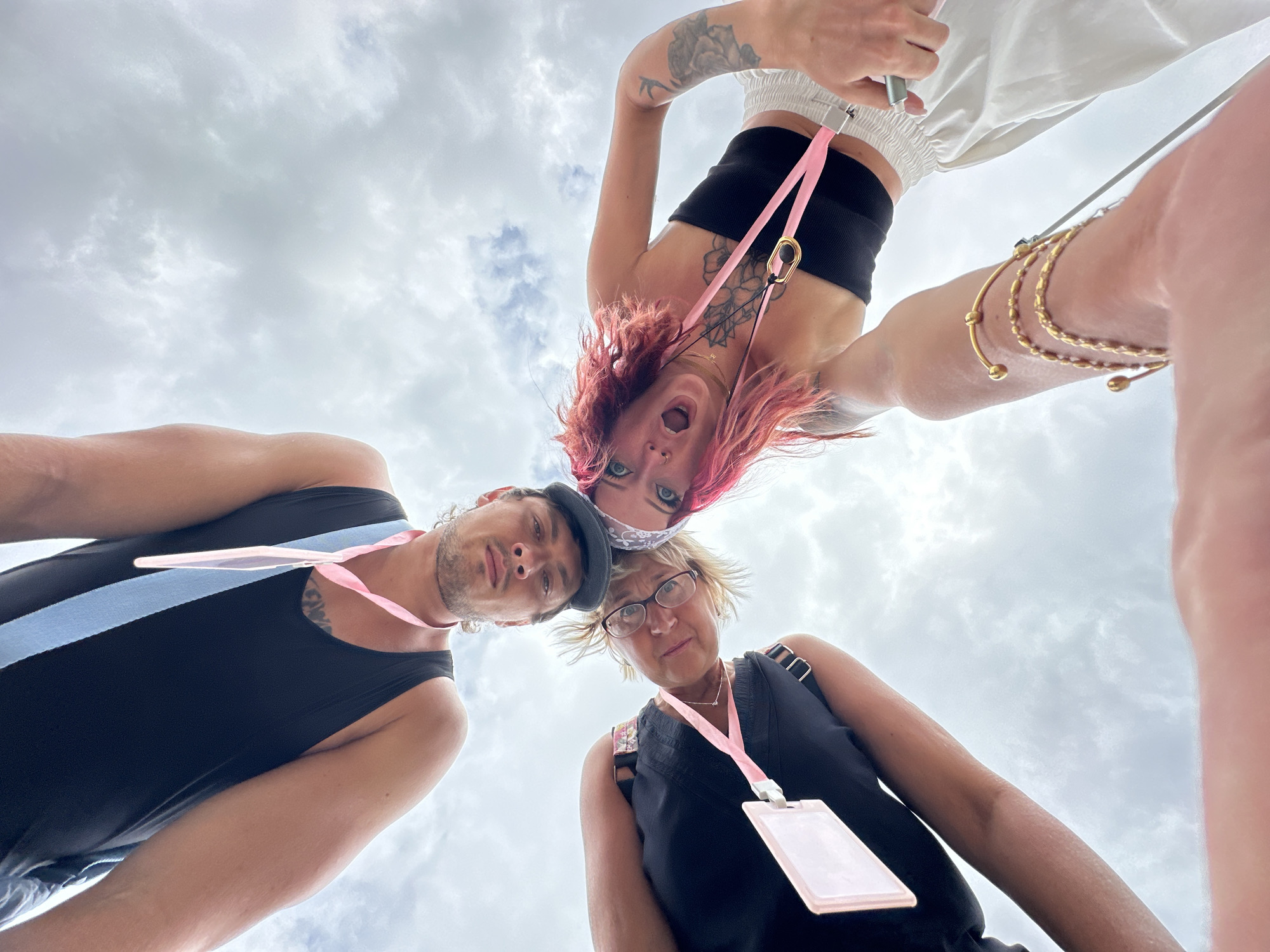 Three people are in the frame, seen from a low angle looking upwards towards a cloudy sky. A man on the left wears a black tank top with a light blue stripe, and a light pink lanyard around his neck. In the middle, a woman with blonde hair and glasses wears a black top with a light pink lanyard holding a badge. On the right, a woman with pink hair and a black top with a light pink lanyard and badge around her neck is looking down at the camera with an open mouth. Her arms are raised; one with gold bracelets. She has tattoos on her arm and chest. All three individuals appear to be looking downwards, towards the camera, with varying expressions. The location is Kampung Toyapakeh, Indonesia.