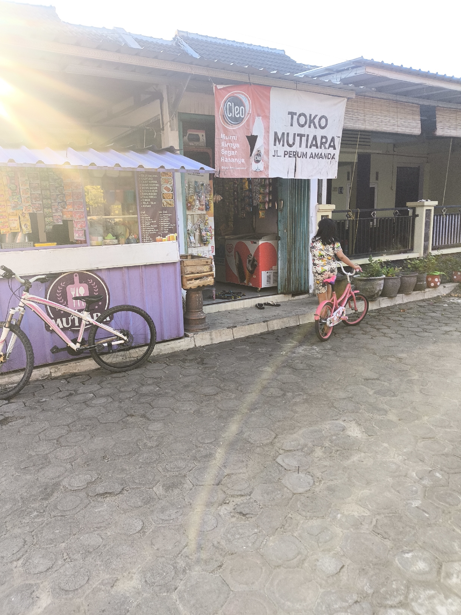 In Lumajang, Indonesia, an outdoor scene captures a street-level view of a small shop and its surroundings. A banner displaying "TOKO MUTIARA" and an address reads "JL. PERUM AMANDA." Adjacent to the shop, a purple stall displays various packaged goods. A pink and white bicycle rests against the purple stall, its front wheel facing left and its rear wheel to the right. Further down the sidewalk, a young girl in a floral-patterned outfit rides a pink bicycle, facing towards the right of the frame. Behind her, potted plants line a fence. An open freezer unit with a red and white design is visible to the left of the girl. The ground is paved with hexagonal concrete blocks. The sun shines brightly, casting lens flare across the upper left portion of the image, suggesting it's a sunny day.