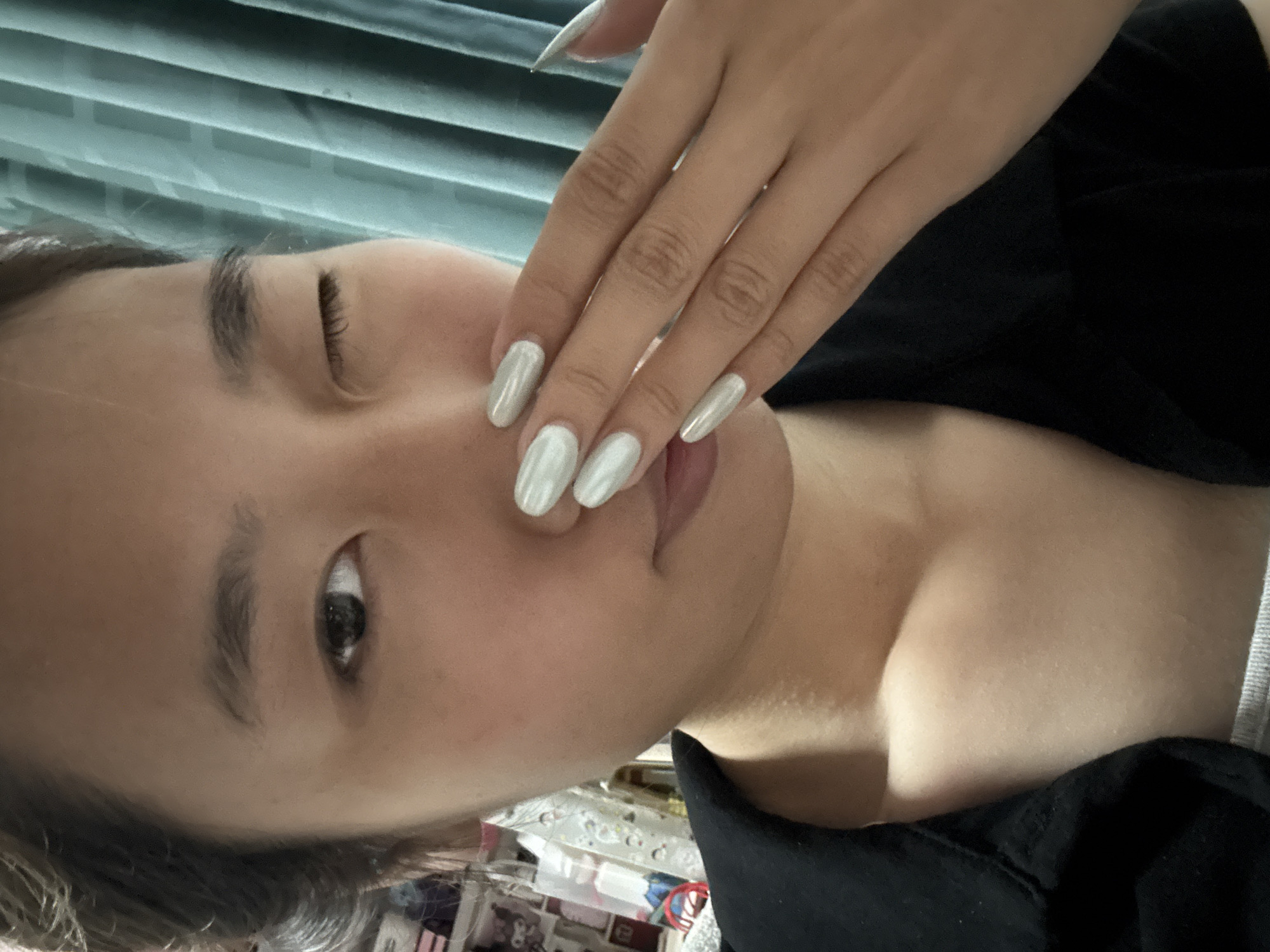 The image shows a person with fair skin and dark hair. The person is facing the camera with one eye open and one closed, suggesting a wink. The person's hand is positioned near their face, showcasing long, almond-shaped nails with a metallic silver polish. The person appears to be wearing a black garment. The background is slightly blurred but shows horizontal lines suggesting blinds or curtains.
While it is impossible to determine the exact location in Nan'an District, China, or any other specific details about the scene from this single image, the visible elements suggest an indoor setting, likely a personal space.