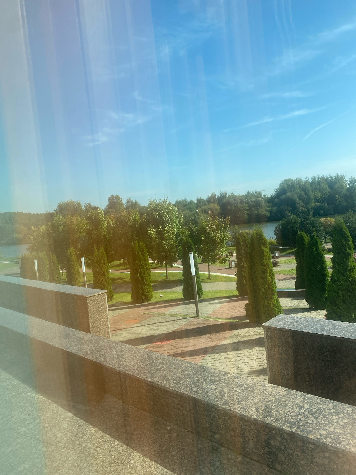 The image displays a sunlit outdoor scene with a clear blue sky and a body of water in the background, suggesting a park or waterfront area. In the foreground, a granite structure with a speckled pattern forms a barrier. Beyond this, a paved walkway with reddish and yellowish bricks leads through a manicured landscape featuring rows of evergreen trees and scattered deciduous trees. Several lamp posts are visible among the trees. In the middle ground, the trees form a dense line, and further back, a calm body of water is visible, bordered by a line of trees and vegetation. The overall impression is one of a peaceful, well-maintained urban outdoor space. The context of Minsk, Belarus, suggests this could be a location along the Svislach River or one of the city's numerous parks.