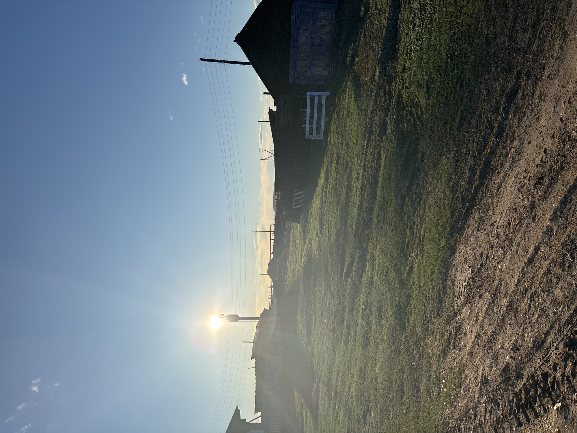The image depicts a rural scene near Zagatuy, Russia. The landscape consists of a grassy field adjacent to a dirt road marked with tire tracks. Several wooden structures, including buildings and fences, are visible, extending towards the horizon. Power lines stretch across the sky. The sun, positioned low in the sky, emits a strong glare and casts long shadows across the landscape.