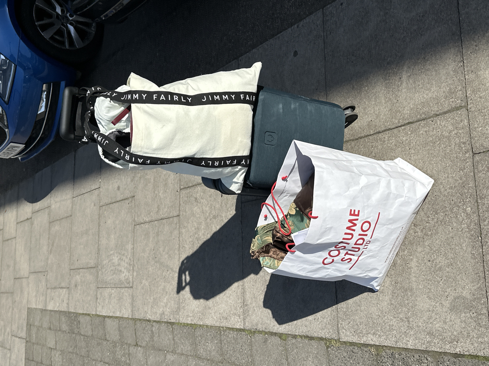 The image captures a street scene in London, United Kingdom, showcasing a luggage set positioned next to a blue car. The luggage includes a teal suitcase with a white tote bag resting on top, marked with the repeated text "JIMMY FAIRLY." Adjacent to the suitcase is a white shopping bag from "COSTUME STUDIO LTD," overflowing with items of fabric. The scene is set on a concrete sidewalk, with the vehicle partially visible on the left side of the frame.