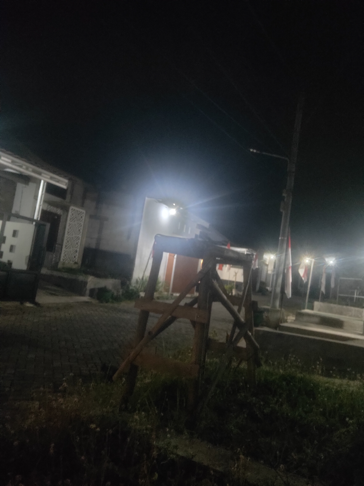 The image depicts an outdoor scene at night, likely in a residential area of Antirogo, Indonesia, based on the presence of Indonesian flags. In the foreground, a wooden A-frame structure is visible, constructed from rough-hewn lumber. The structure appears to be a temporary scaffold or support. To the left of the frame, a paved walkway or driveway is present, bordered by overgrown grass and vegetation.
In the background, several buildings are discernible. One building on the left has a modern facade with clean lines and recessed lighting. Adjacent to it is another building with decorative geometric patterns on its exterior wall. Further into the background, another building with a lighter facade is illuminated by bright lights, creating a glare. A utility pole with power lines is also visible, supporting a street lamp. Several Indonesian flags, white and red, are displayed on poles, some appearing to be attached to the utility pole. The overall environment is dark, with artificial lighting from buildings and street lamps illuminating sections of the scene. No people are visible in the image.