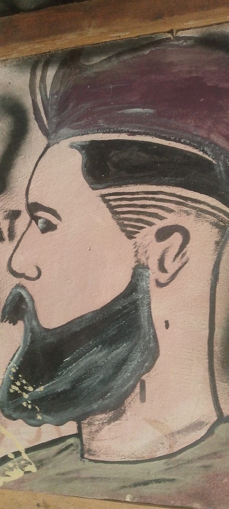 A painted portrait of a man's head and shoulders is depicted. The man has a dark, full beard and mustache. His hair is styled with a side part and appears to be slicked back. He is wearing a dark garment on his shoulders. The background is a textured, light pinkish-brown color. The portrait appears to be framed by a wooden border. The style is illustrative and appears to be a mural or wall art. The location of this artwork in Jalingo, Nigeria, is not provided in the image itself.