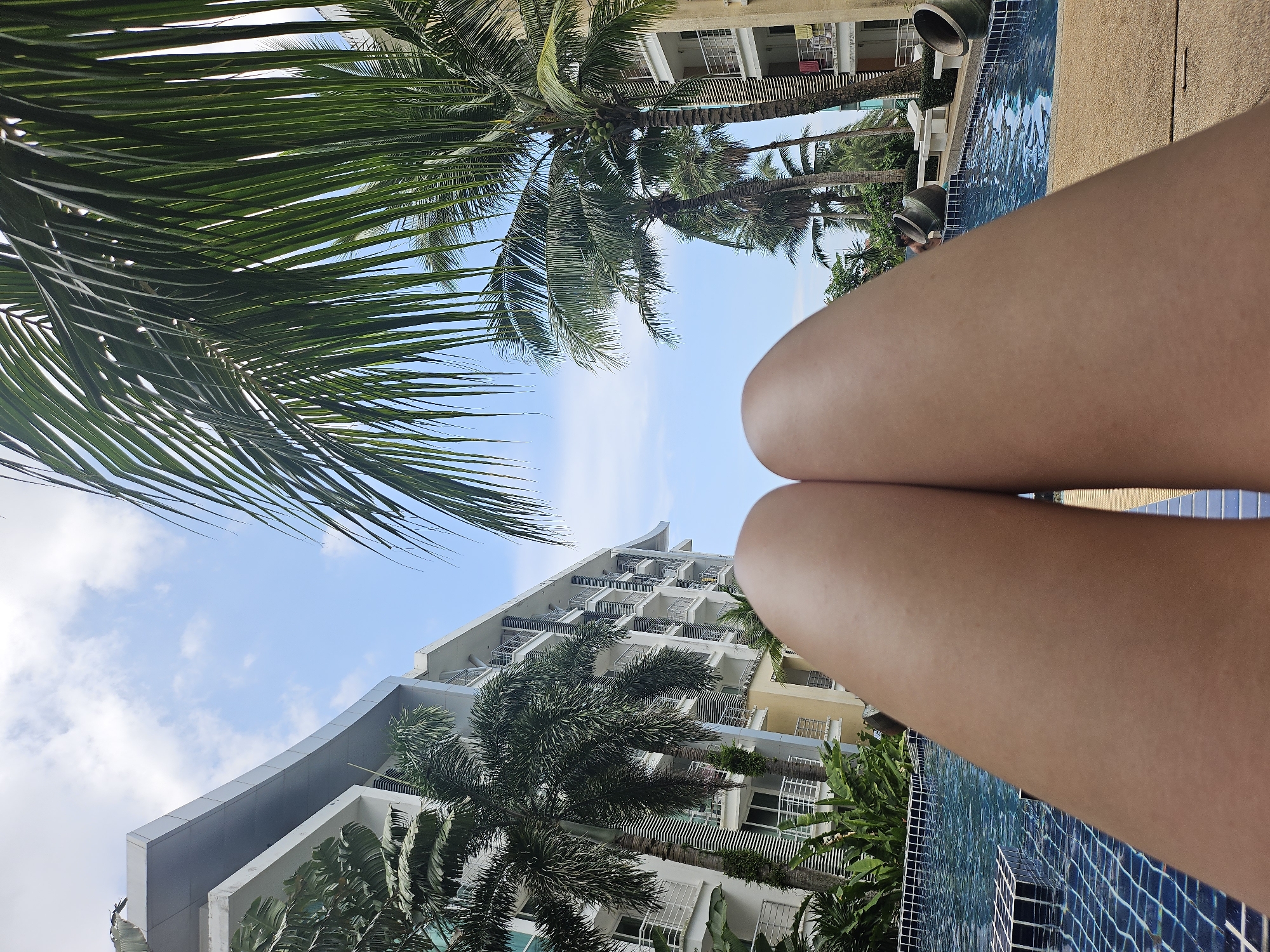 The image depicts a first-person perspective of a person relaxing by a swimming pool, with their legs prominently in the foreground. The setting appears to be a resort or hotel in Kathu, Thailand, evidenced by the presence of palm trees, modern architecture resembling resort buildings, and the overall tropical ambiance. The swimming pool is tiled in blue and appears clean. In the background, there are several multi-story buildings with balconies, suggestive of hotel rooms or apartments. Palm trees with lush green fronds are scattered around the pool area and between the buildings, creating a tropical atmosphere. The sky is blue with scattered white clouds.
