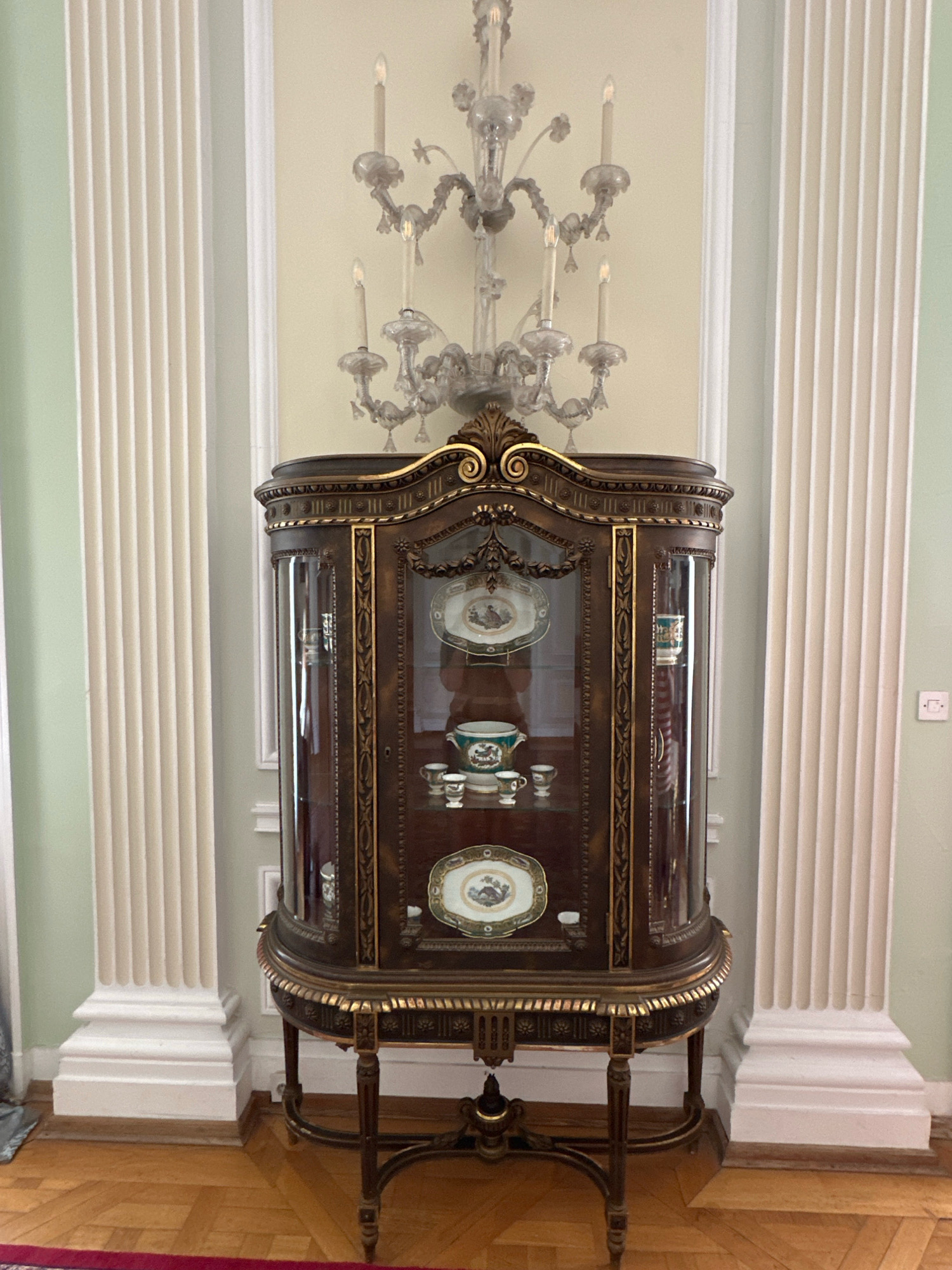 The image shows an ornate, dark wood display cabinet with glass doors, filled with porcelain dishes and teacups. The cabinet is ornately decorated with gold trim and rests on a carved wooden stand. Above the cabinet, a crystal chandelier with several candle-shaped lights is visible. The cabinet is positioned in front of a pale green wall, flanked by two large, fluted white columns. The floor is made of polished light-colored wood arranged in a parquet pattern. This scene is likely from the Royal Palace in Belgrade, Serbia, a location known for its historical and ornate furnishings. There are no people visible in the image, and the objects are stationary.