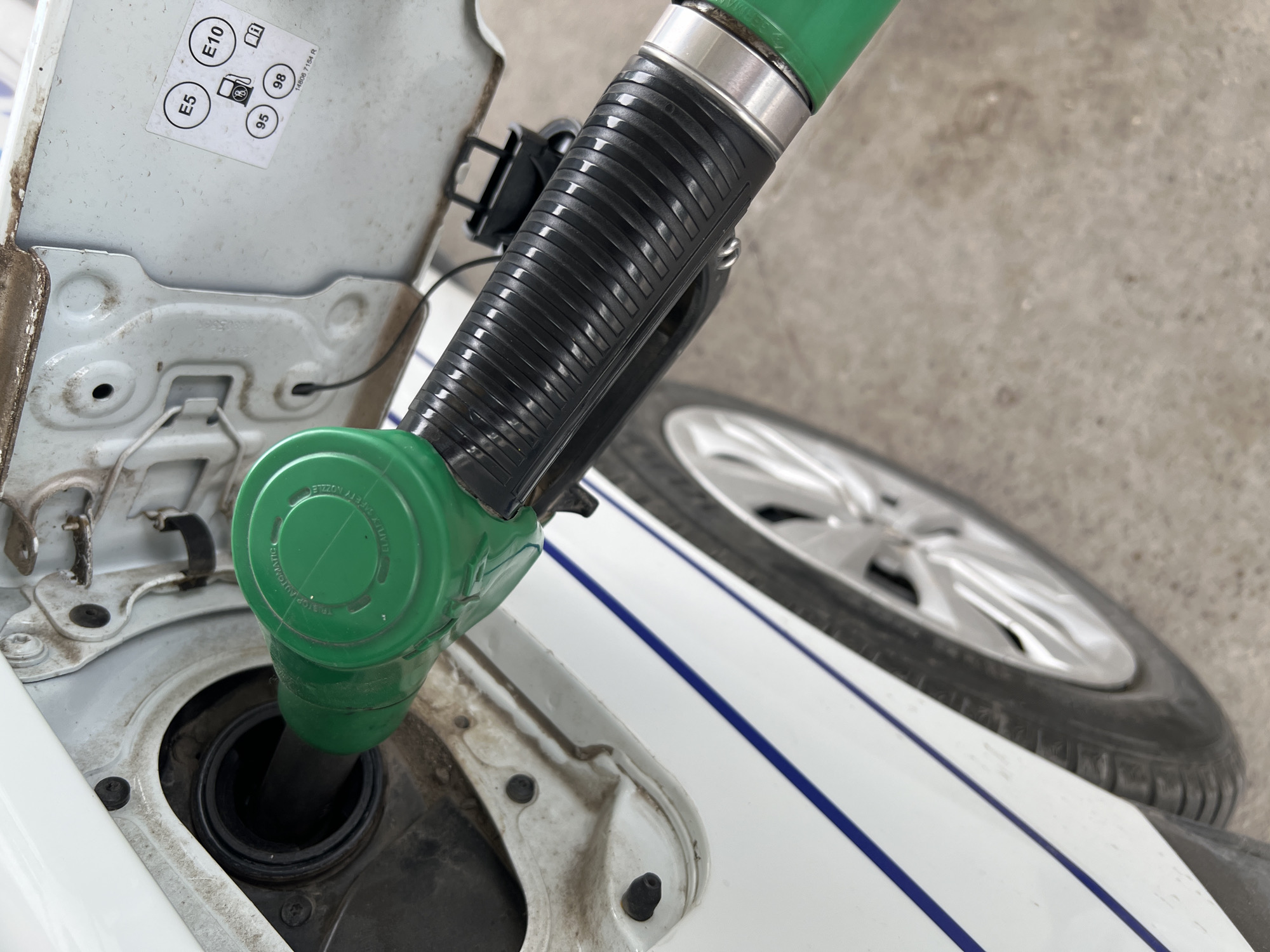A close-up, high-angle shot captures a green fuel pump nozzle inserted into the fuel tank of a white car. The car's fuel flap is open, revealing the fuel filler neck and the surrounding bodywork, which shows some dirt and grime.
On the inside of the fuel flap, a white sticker with black text is clearly visible. It displays fuel types and octane ratings: "E5", "E10", "95", and "98". There is also a circular icon depicting a fuel pump with a letter "B" inside, and another icon representing an owner's manual. A serial or part number "14806 7154 R" is also printed on the sticker.
The green nozzle has a black ribbed grip and a metallic silver collar. Faint text can be seen on the green body of the nozzle, including phrases like "TO STOP AUTOMATICALLY" and "SAFETY NOZZLE".
In the background, part of the car's front or rear wheel, with a silver alloy rim and black tire, is visible. The ground appears to be concrete. The white car body features a thin blue stripe running horizontally along its side. The scene depicts the common act of refueling a vehicle at what appears to be a gas station.
