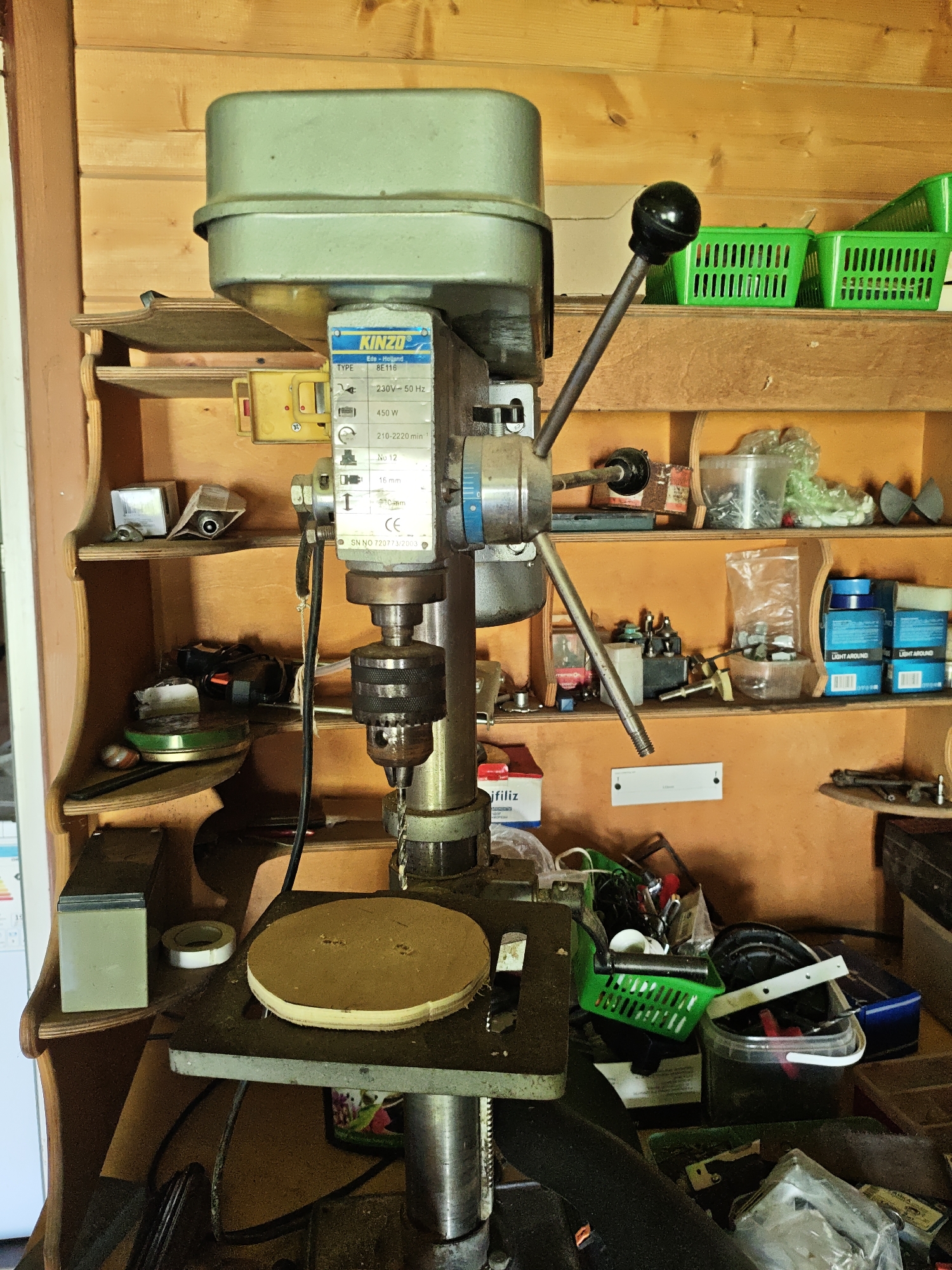 The image shows a Kinzo drill press in what appears to be a cluttered workshop setting. The drill press, model BE116, is light green and silver with a black handle. A round piece of wood is clamped on the drill press table. Behind the drill press are wooden shelves holding various tools, supplies, and containers. The background is wooden paneling. The OCR data includes "Ede-Holland," the Kinzo brand, and specifications like voltage, wattage, and speed, alongside a CE mark and serial number. No people are visible in the image. There is no information related to the location in the city of Боровково, Russia.