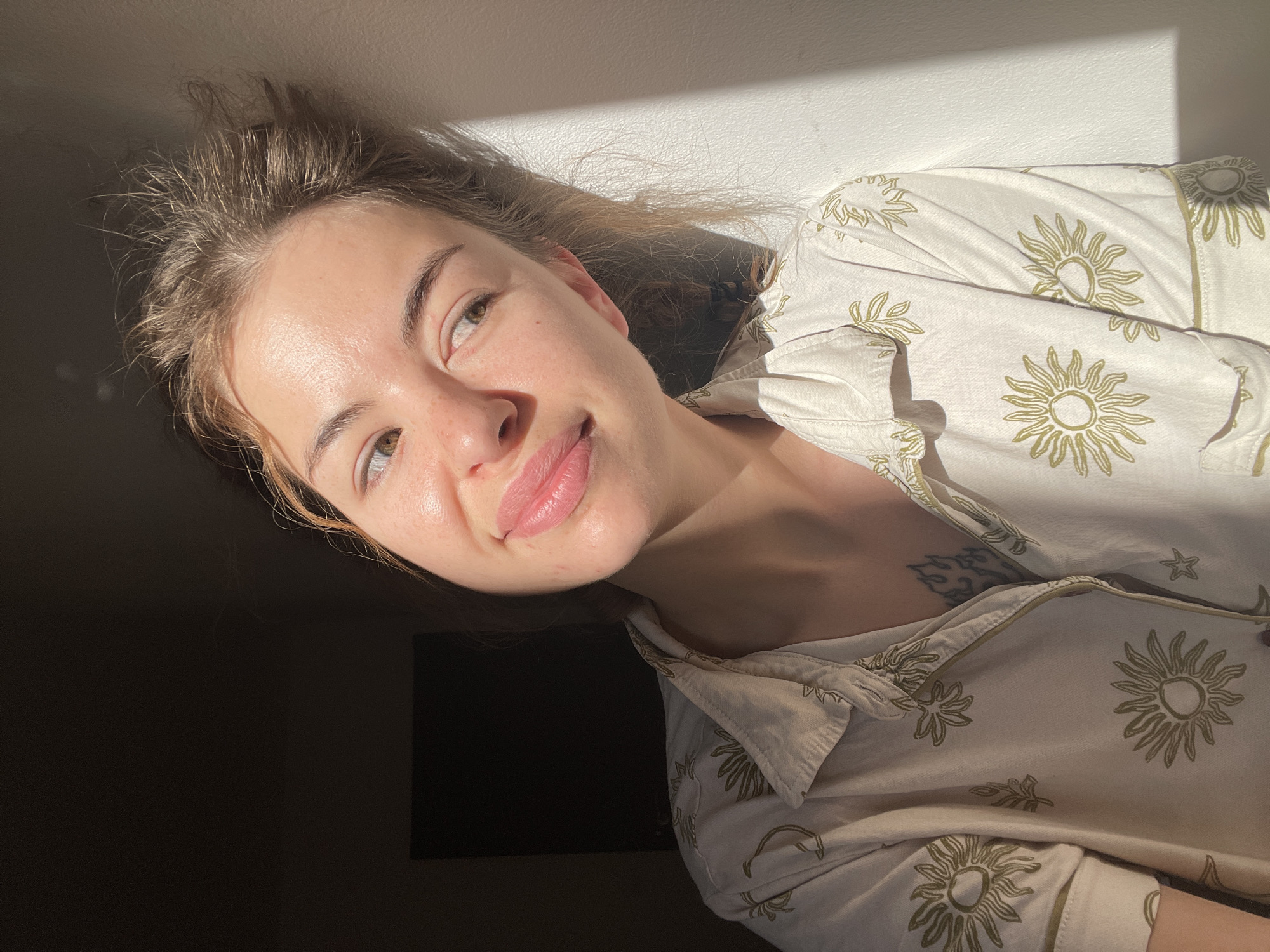 A woman is facing the camera. She has light brown hair and fair skin. She is wearing a light-colored button-down shirt with an olive-green sun pattern on it, and there is a tattoo visible on her upper chest. The lighting is dramatic, with strong sunlight creating highlights and shadows on her face and the wall behind her. The background is partially obscured by darkness. There is no discernible evidence that the location is Boreti, Montenegro.
