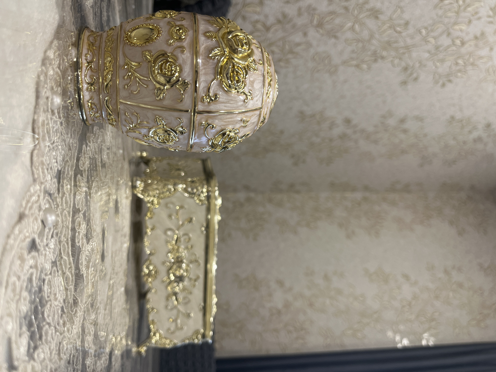 The image shows two ornate decorative boxes on a reflective surface, possibly a table. The box in the foreground is egg-shaped, with a pinkish hue and intricate gold floral embellishments, including roses. It has a base with decorative trim. In the background, slightly out of focus, is a rectangular box, also cream-colored with gold decorative elements and ornate legs. The backdrop is a textured wallpaper with a subtle floral pattern. The lighting suggests an indoor setting, likely a room in a residential property. The scene is static and does not depict any people or actions. The location, Никольское, Russia, is mentioned but cannot be visually confirmed from the image itself.