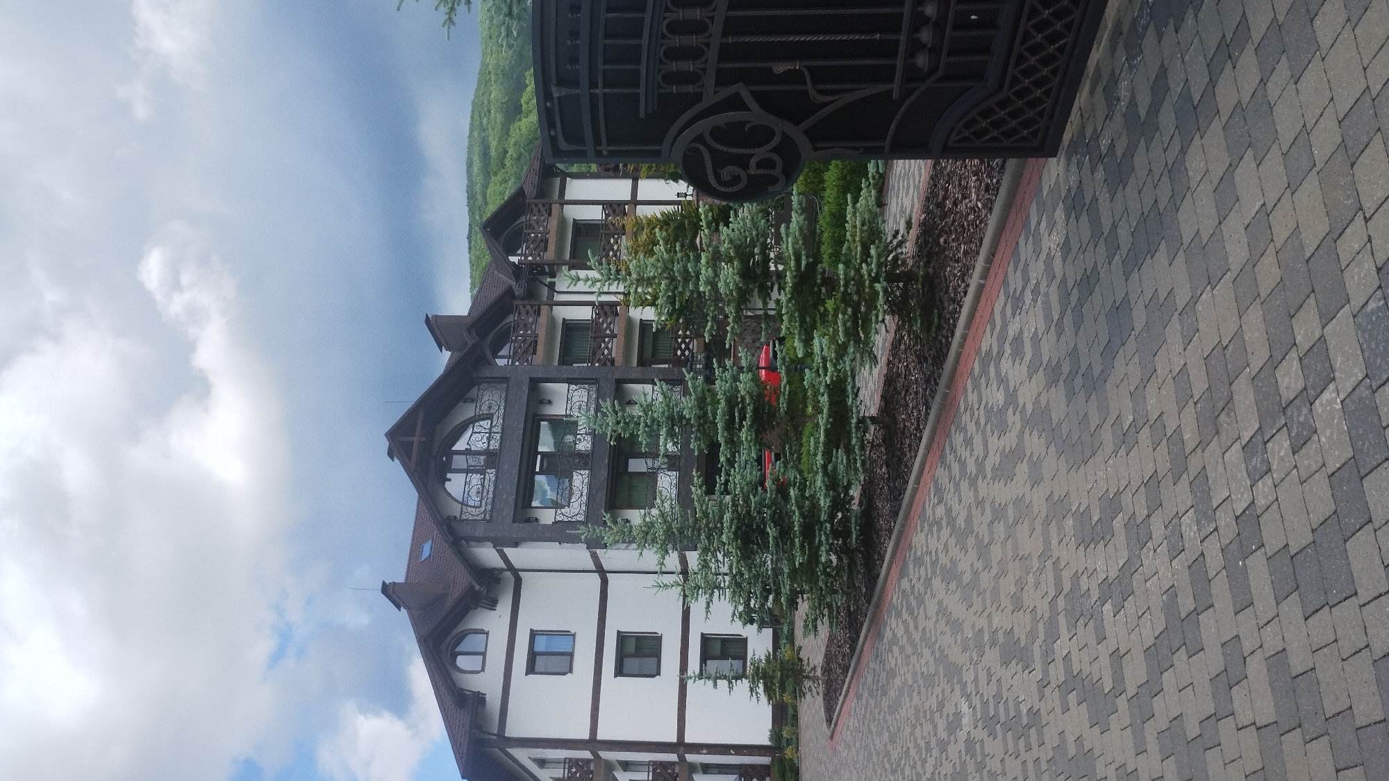 The image shows a multi-story building with a dark roof and white and dark grey facade, lined with small evergreen trees along a paved driveway. A large, ornate black metal gate is partially open on the right side of the frame. The building has several balconies with wooden railings and decorative metalwork. In the background, a lush green hillside is visible under a cloudy sky. The scene appears to be a residential or hotel property, possibly located in the city of Syniak, Ukraine.