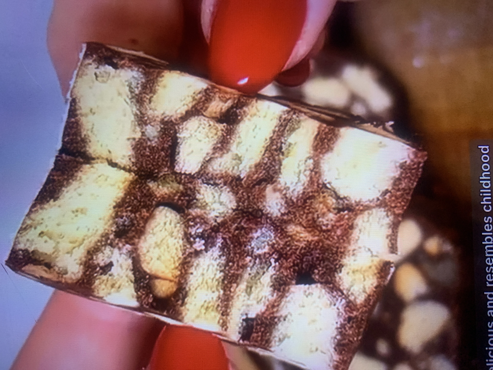 A slice of chocolate biscuit cake is held by hands with red nail polish. The cake has layers of biscuit pieces and chocolate. The background is out of focus. The image is a close-up of the cake and hands. There is text at the bottom that says "delicious and resembles childhood". No location in Almaty, Kazakhstan is visible.