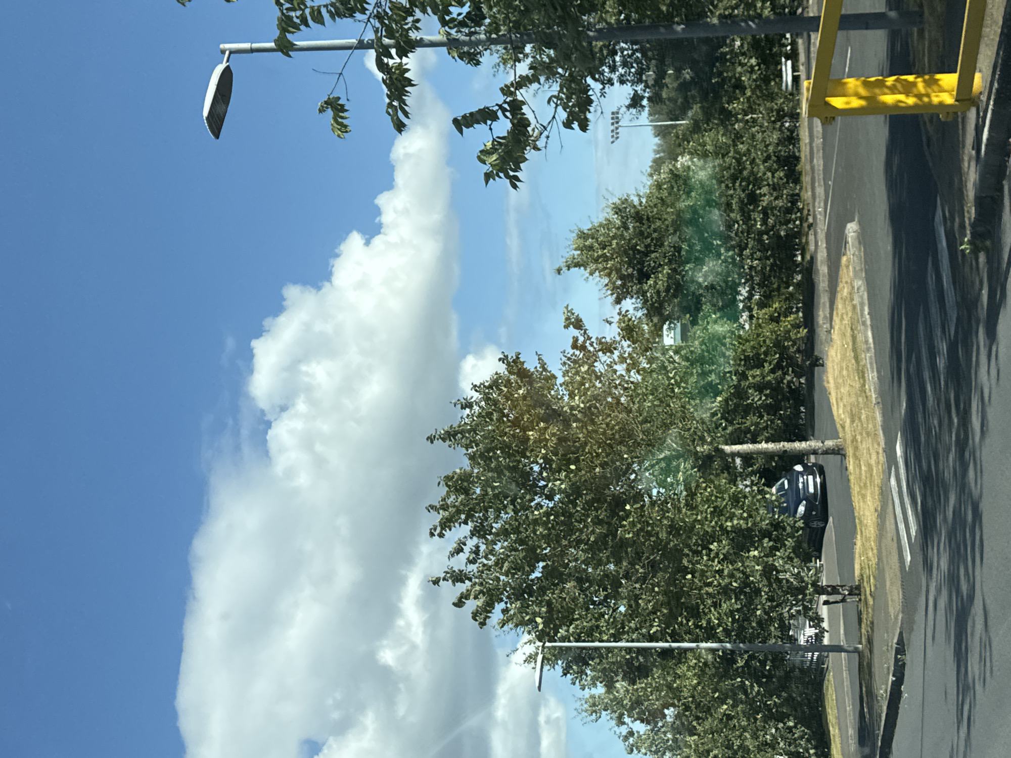 A dark sedan is parked on the right side of a paved lot, partially obscured by trees and bushes. A yellow bollard stands in the foreground on the right. The sky is blue with scattered white clouds, and the sun is shining. Trees and streetlights are visible throughout the scene. The location is the city of Waterford, Ireland.