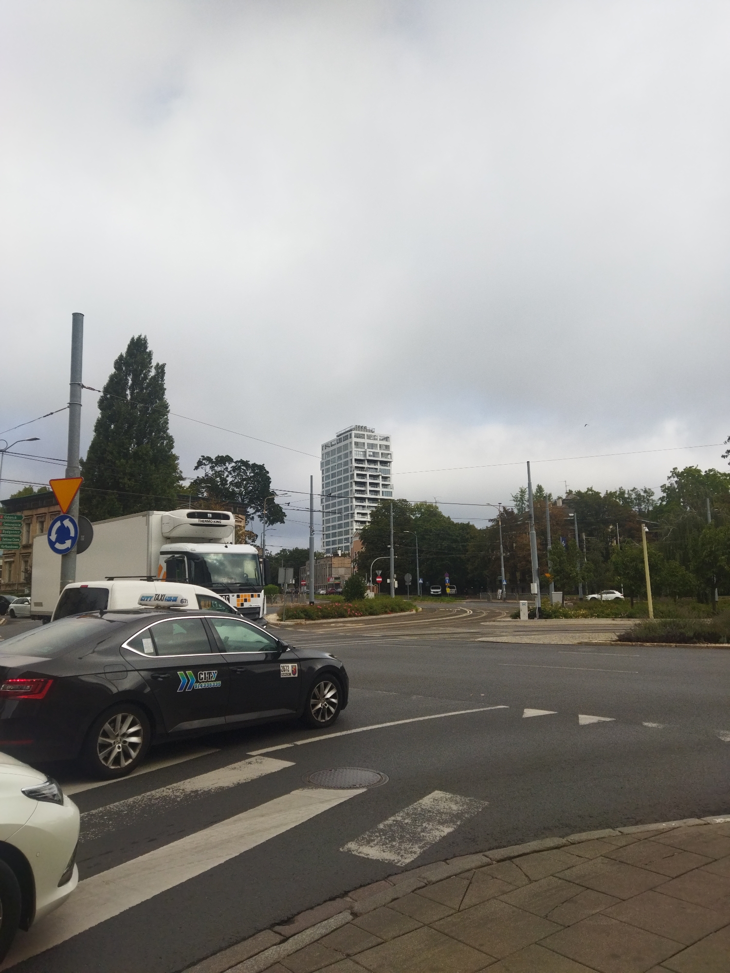 A wide shot captures a city intersection on a cloudy day. A dark sedan, identified as a "CITY TAXI" by its livery, is parked in the foreground, partially obscuring a white car. Behind the taxi, a white truck with a "THERMO KING" logo is stopped at the intersection. A multi-story building with a modern, glass facade stands tall in the background, towering over a dense cluster of trees. Traffic lights and street signs are visible, indicating the intersection is active. The road surface shows clear lane markings and pedestrian crossings. The image is likely taken in Szczecin, Poland, as suggested by the presence of a directional sign indicating cities like Gdansk, Swinoujscie, and Kolobrzeg.