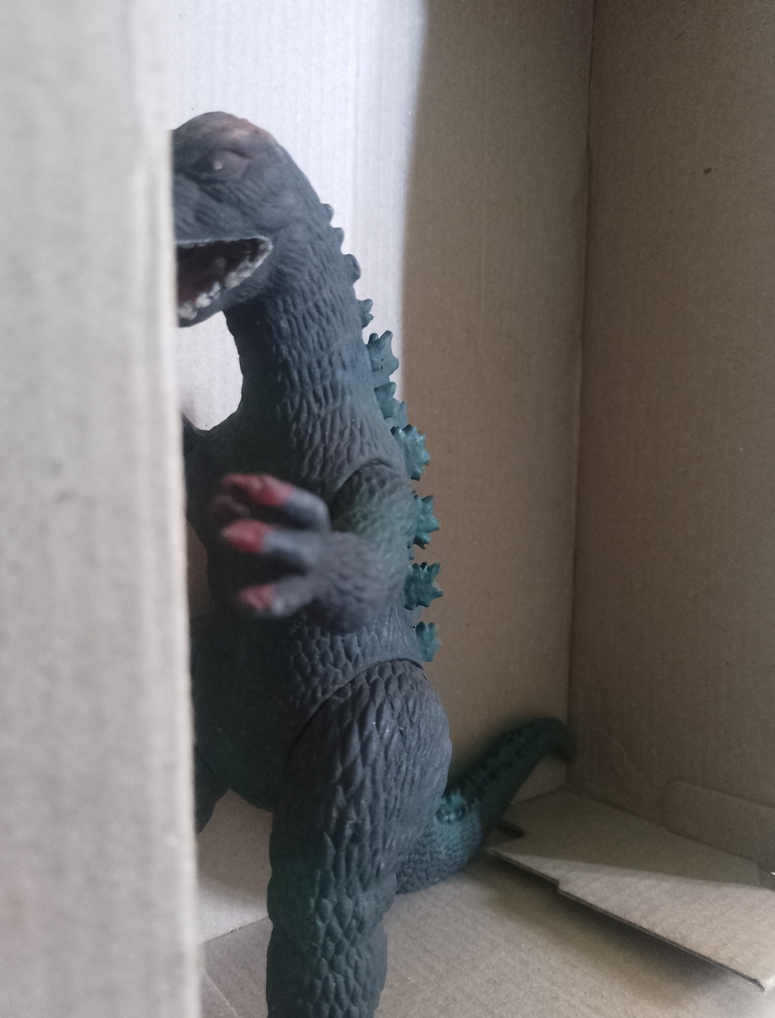 A dark green, toy Godzilla figure is standing in a cardboard box. The figure is facing right, with its mouth open and its arms extended forward. The figure has red painted claws. The background is the inside of the cardboard box, which is brown. There is no indication of the location in the city of San Lucas Tulcingo, Mexico.