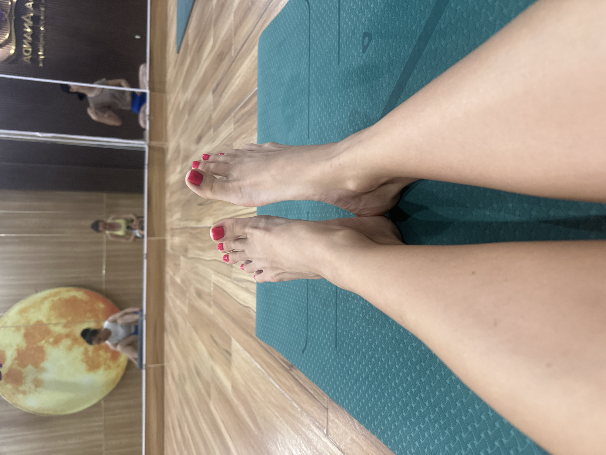 A medium shot shows a pair of female legs and feet, placed on a green yoga mat. The toes are painted with red nail polish. The setting appears to be a yoga studio. Reflections in the mirrored wall reveal a man doing exercises and a yellow moon-like decoration. The floor is wooden. This image was taken in Bac Nha Trang Ward, Vietnam.